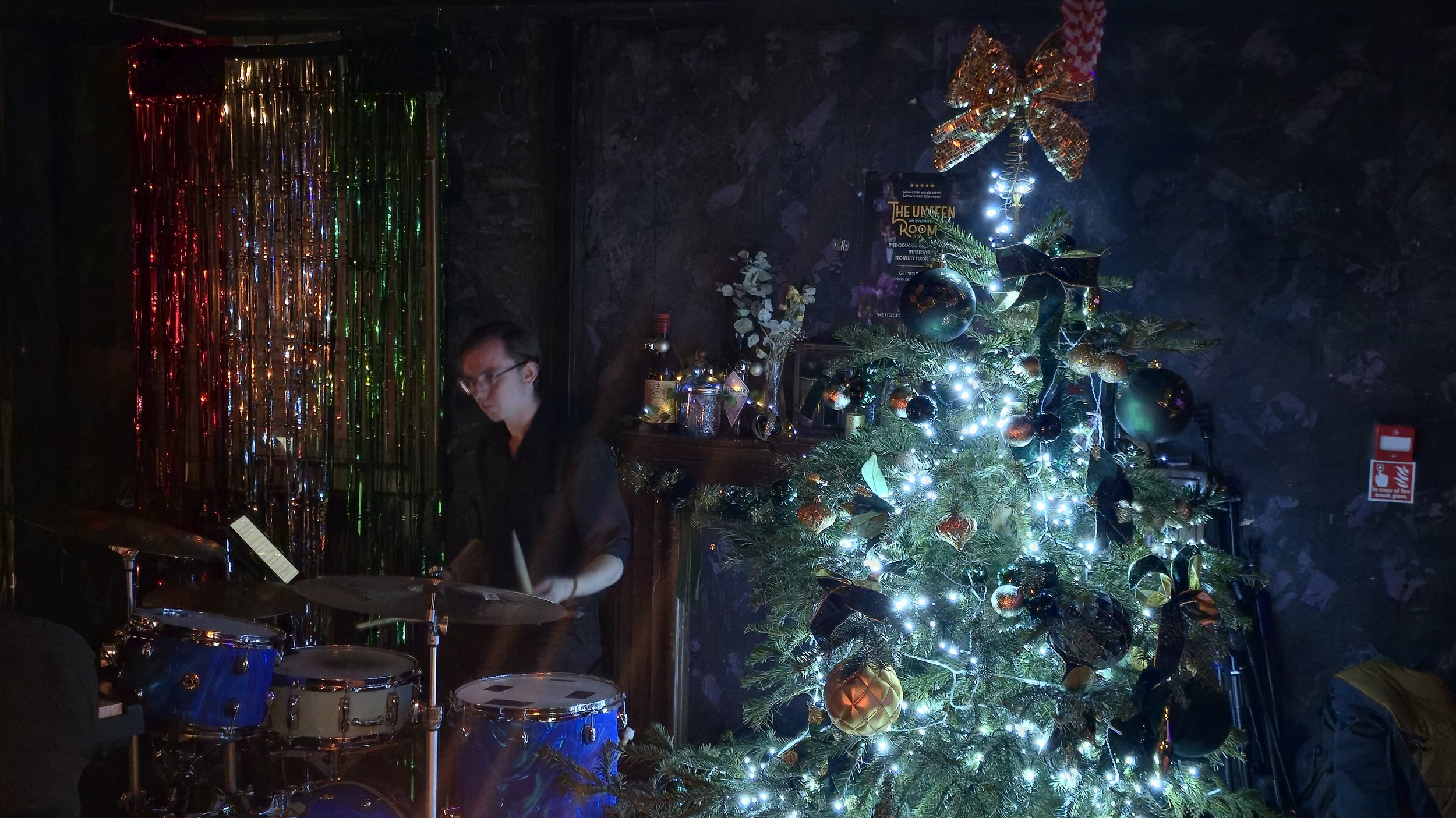 A person playing drums next to a decorated Christmas tree with ornaments and lights, in a dimly lit room with colorful curtain decorations in the background.