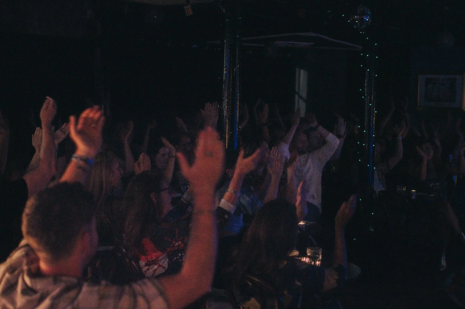 People dancing with hands raised in a dark indoor club or concert venue with colorful lighting and disco ball.