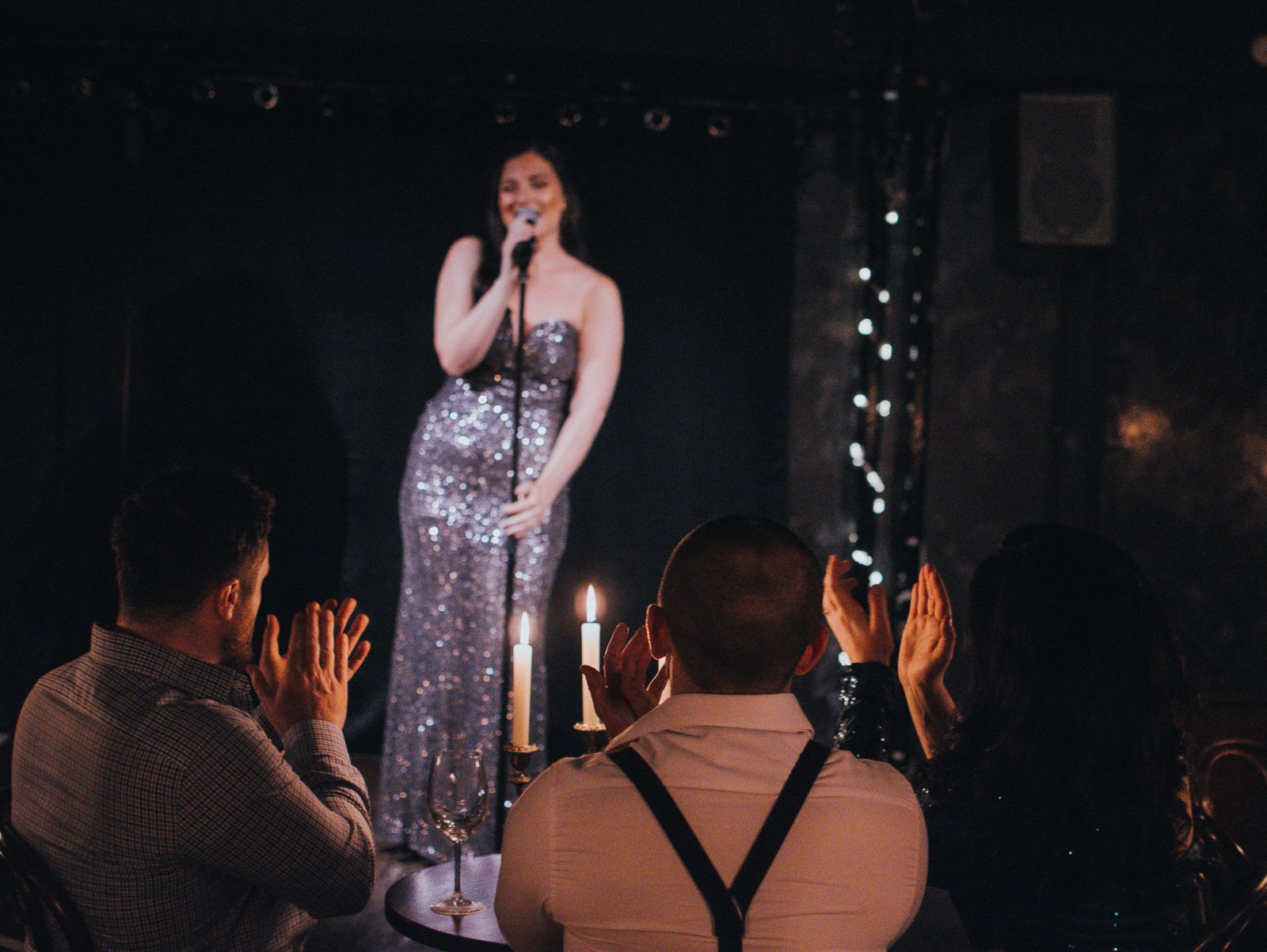 A woman in a sequined dress singing on stage with audience clapping and candles on the table.