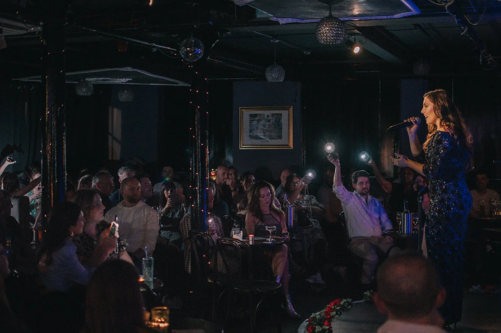 A singer performing on stage with a microphone in hand in a dark venue, with an audience seated at tables watching her. The audience members are holding up their phones and flashlights to watch the performance. The stage is decorated with disco balls and string lights.