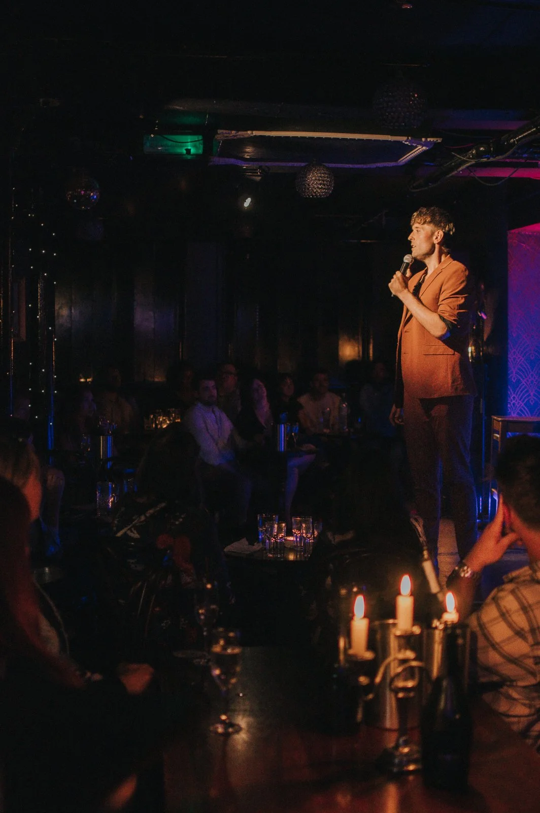 A man in a brown suit performs stand-up comedy on stage holding a microphone, with an audience seated in a dimly lit venue with candles and candles on tables.