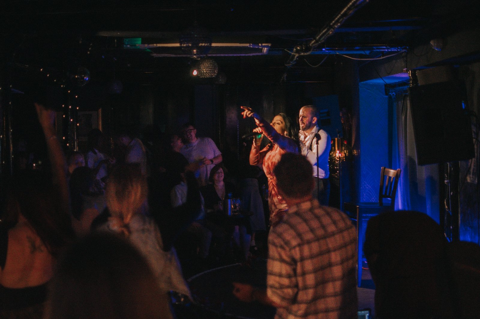 A singer performs on stage with a microphone, accompanied by a guitarist, in a dimly lit bar or nightclub, with audience members watching.