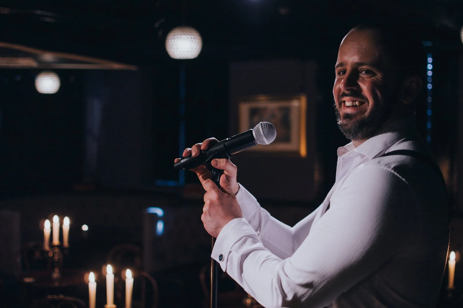 A man with a beard in a white shirt holding a microphone and smiling, standing in a dimly lit venue with candles and framed pictures on the wall.