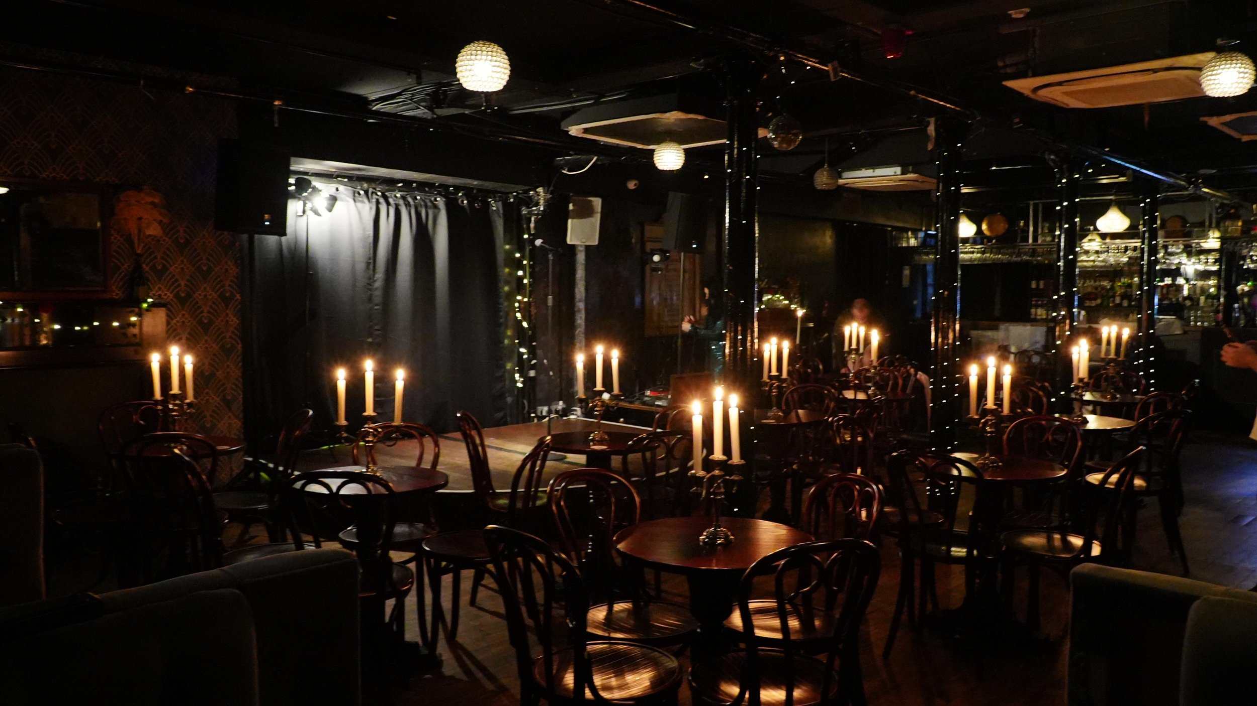 A dimly lit restaurant or bar with tables and chairs, candlelights on the tables, black curtains, and decorative lighting fixtures on the ceiling.