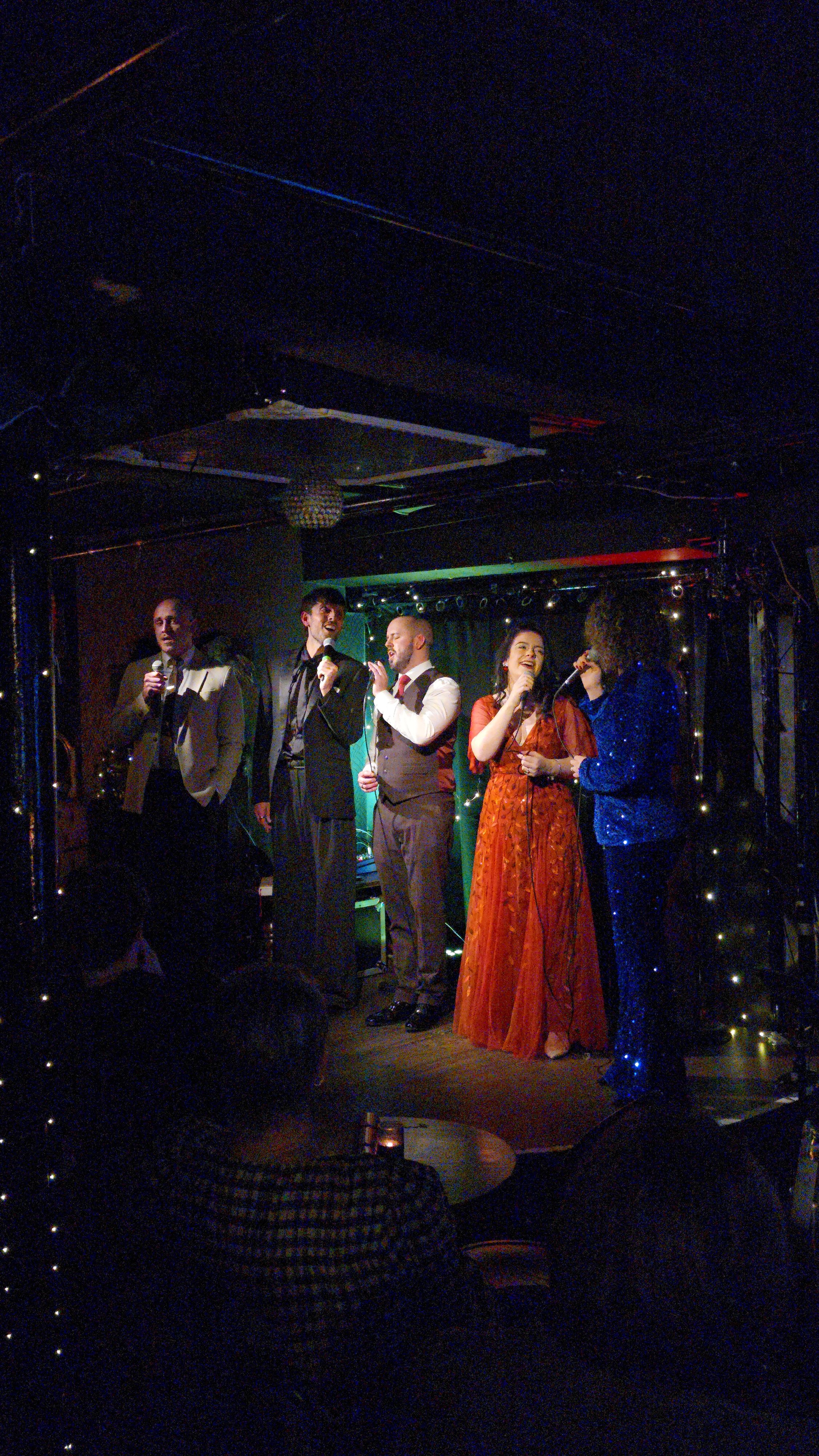Group of five people singing into microphones on stage during a performance in a dimly lit venue, with a woman in a long orange dress and four men in suits.