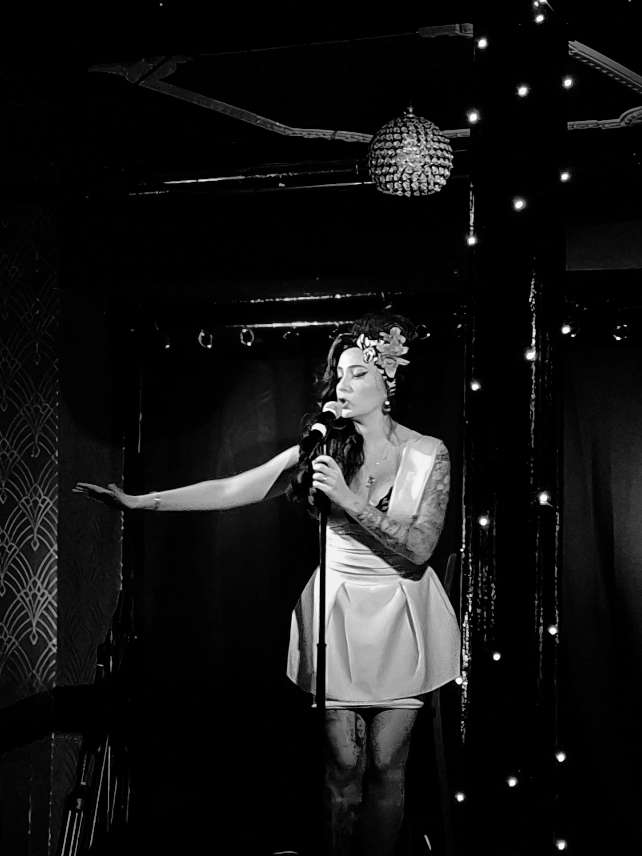 A woman singing into a microphone on stage, wearing a sash and floral headpiece with lights and decorative elements behind her.