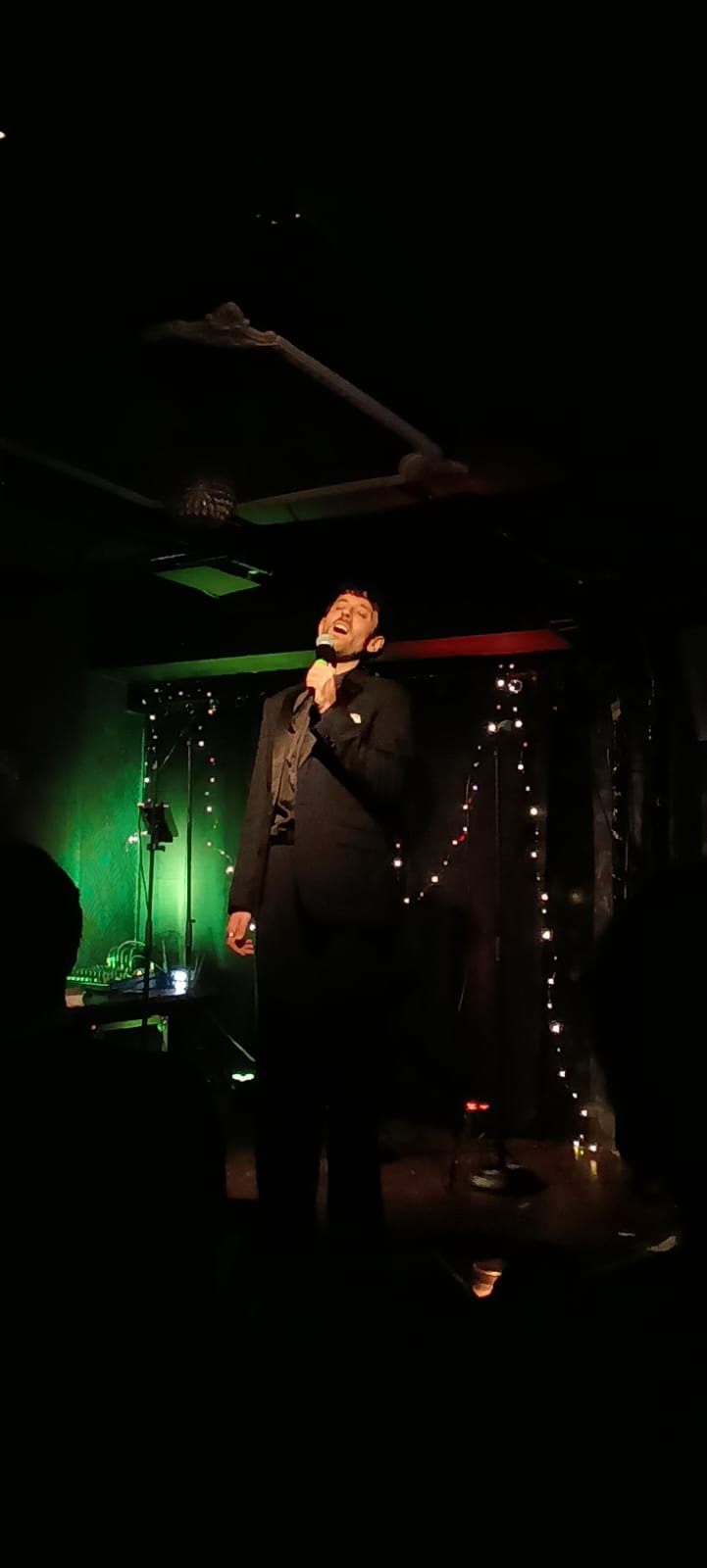 A man in a black suit performing on a stage with a microphone, decorated with string lights, in a dimly lit venue.