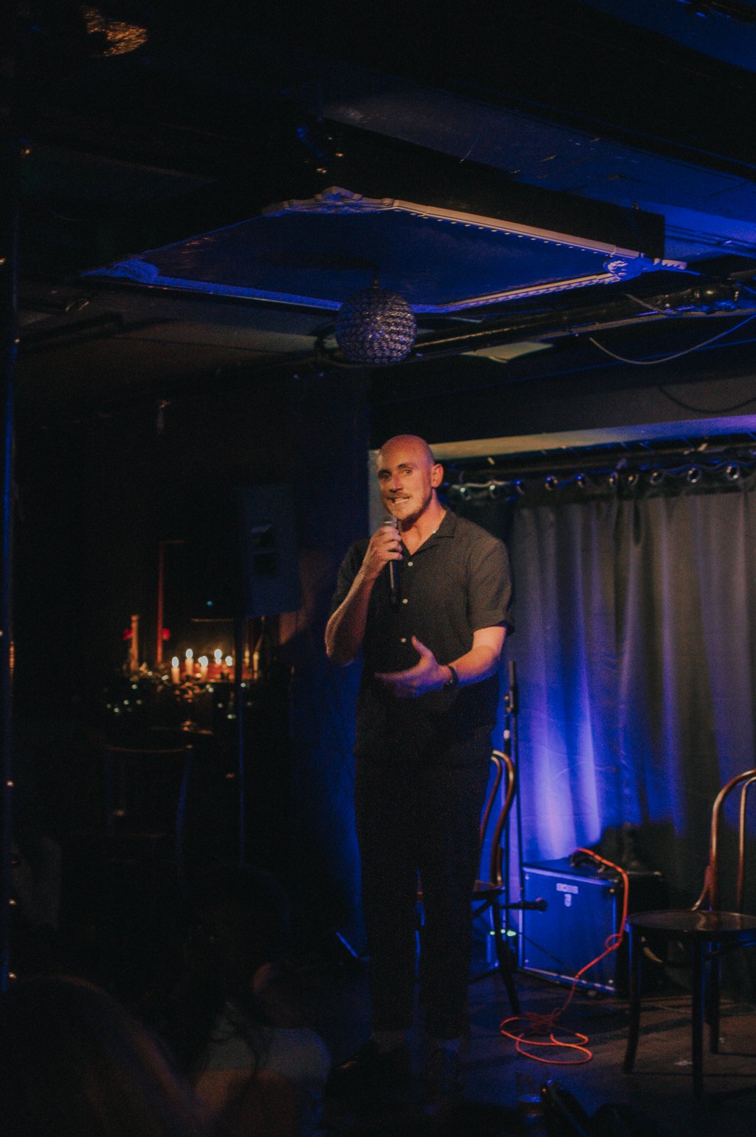 A man with a microphone on stage, smiling, with dark curtains behind him, in a dimly lit entertainment venue.