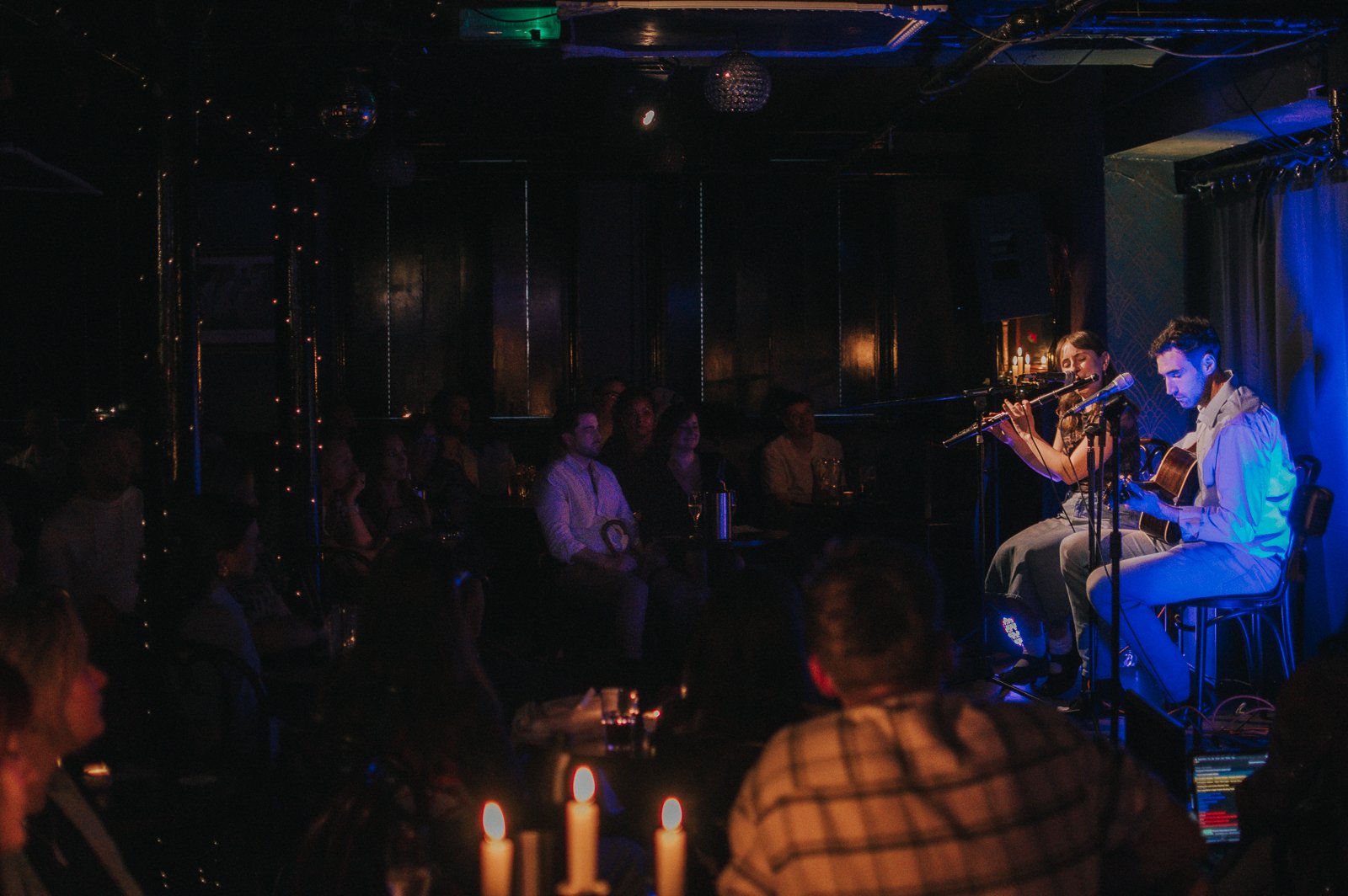 A live music performance at a small venue with an audience watching two musicians, one playing guitar and the other singing or playing flute, on a dimly lit stage.