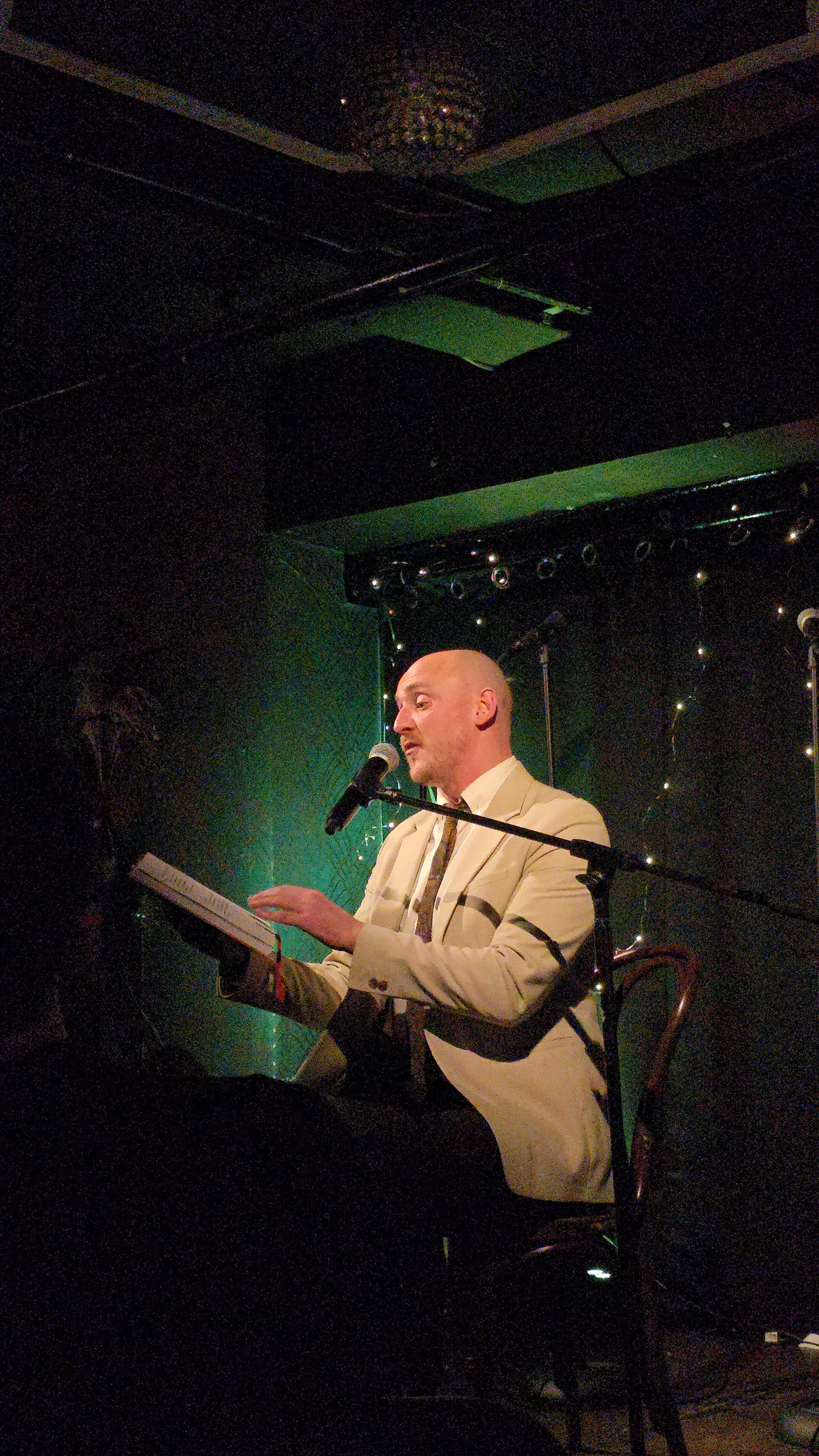 Man in a beige suit performing on stage with a microphone and keyboard in a dimly lit venue.