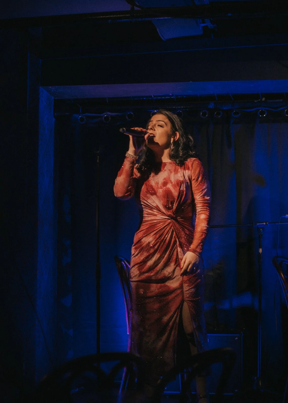 A woman singing on stage with a microphone, wearing a fitted, long-sleeved, red and orange dress with a slit. She has dark, wavy hair and is illuminated by stage lights.