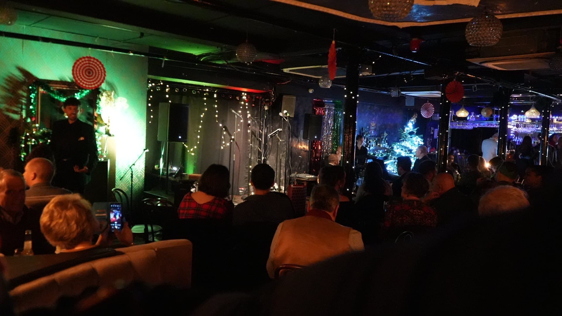 A crowded indoor holiday party or event with people seated at tables, a performer or speaker on stage, and festive Christmas decorations including a decorated tree, wreaths, and Christmas lights.