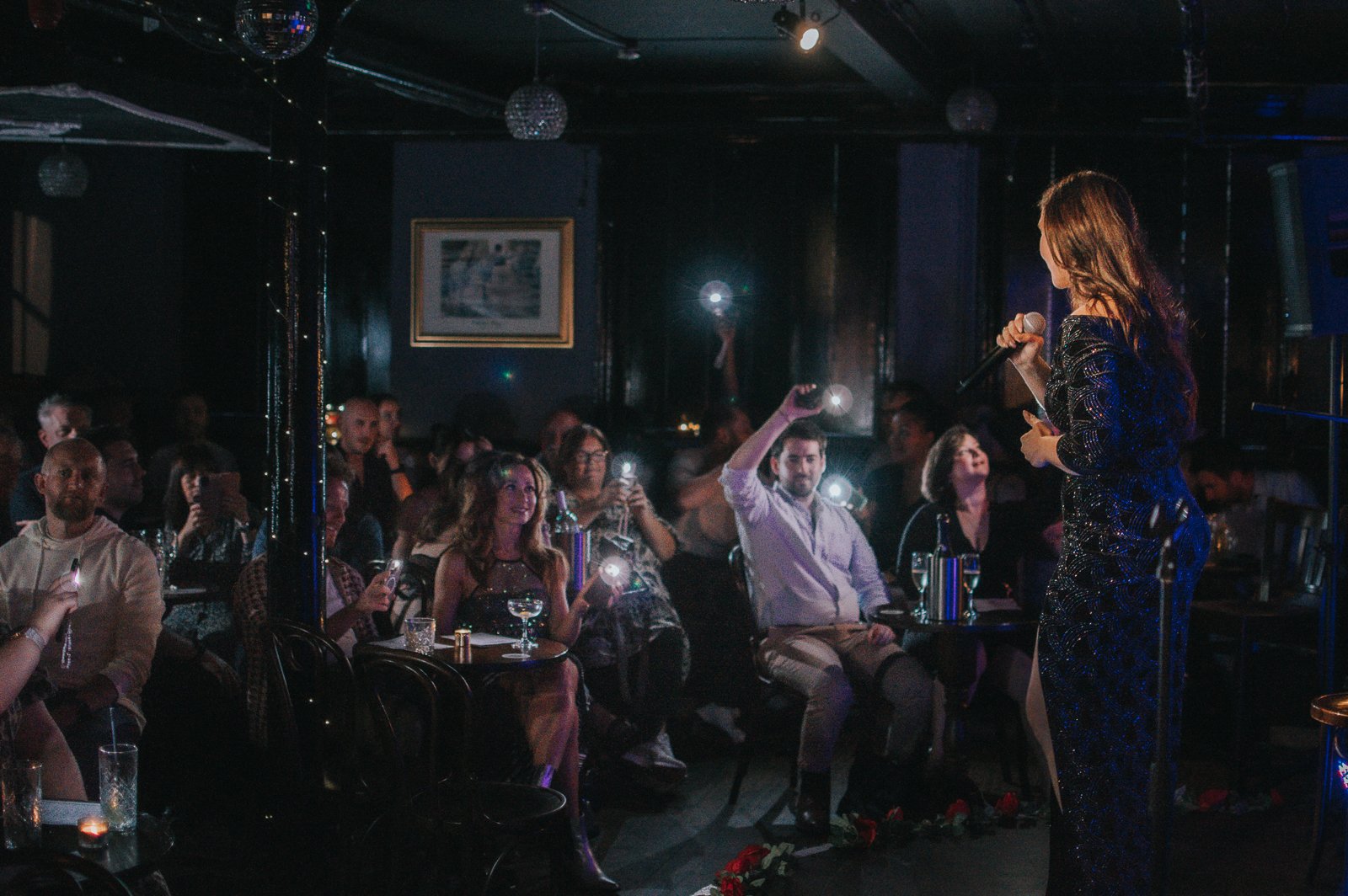 A woman performing karaoke on stage with an audience holding up flashing phones and watching her in a dimly lit venue.