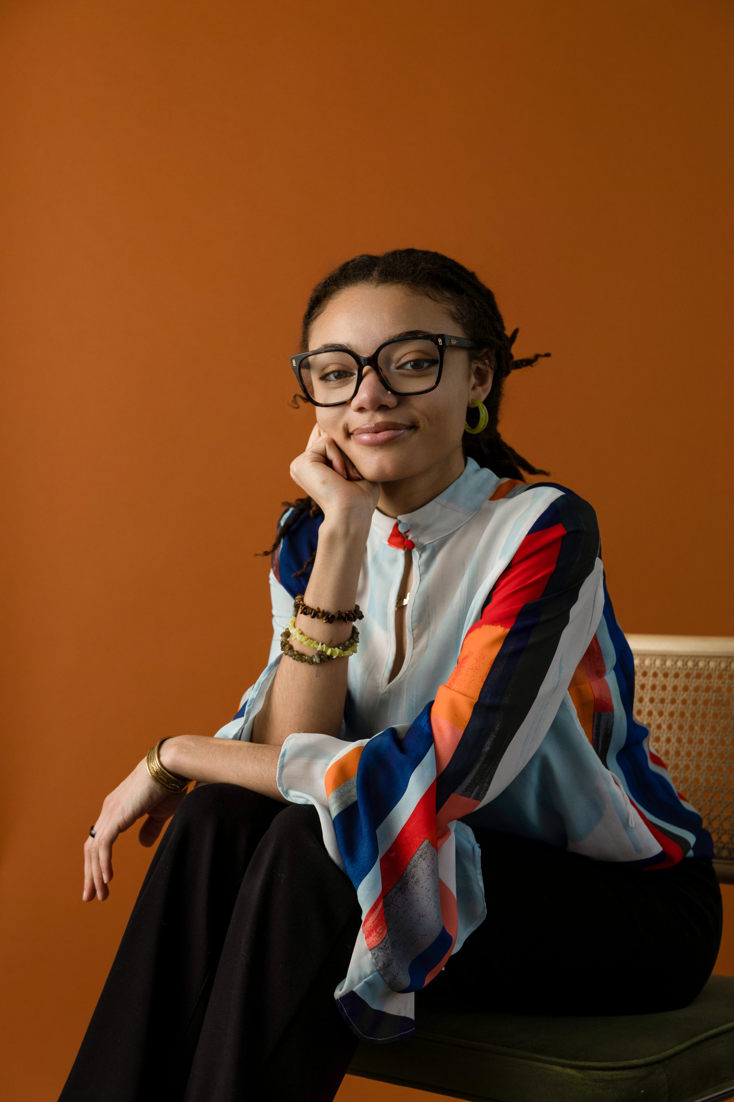London B (LBL) , glasses, and earrings, sitting on a green chair with her chin resting on her hand, against a warm orange background. She is wearing a colorful striped blouse, black pants, and jewelry.