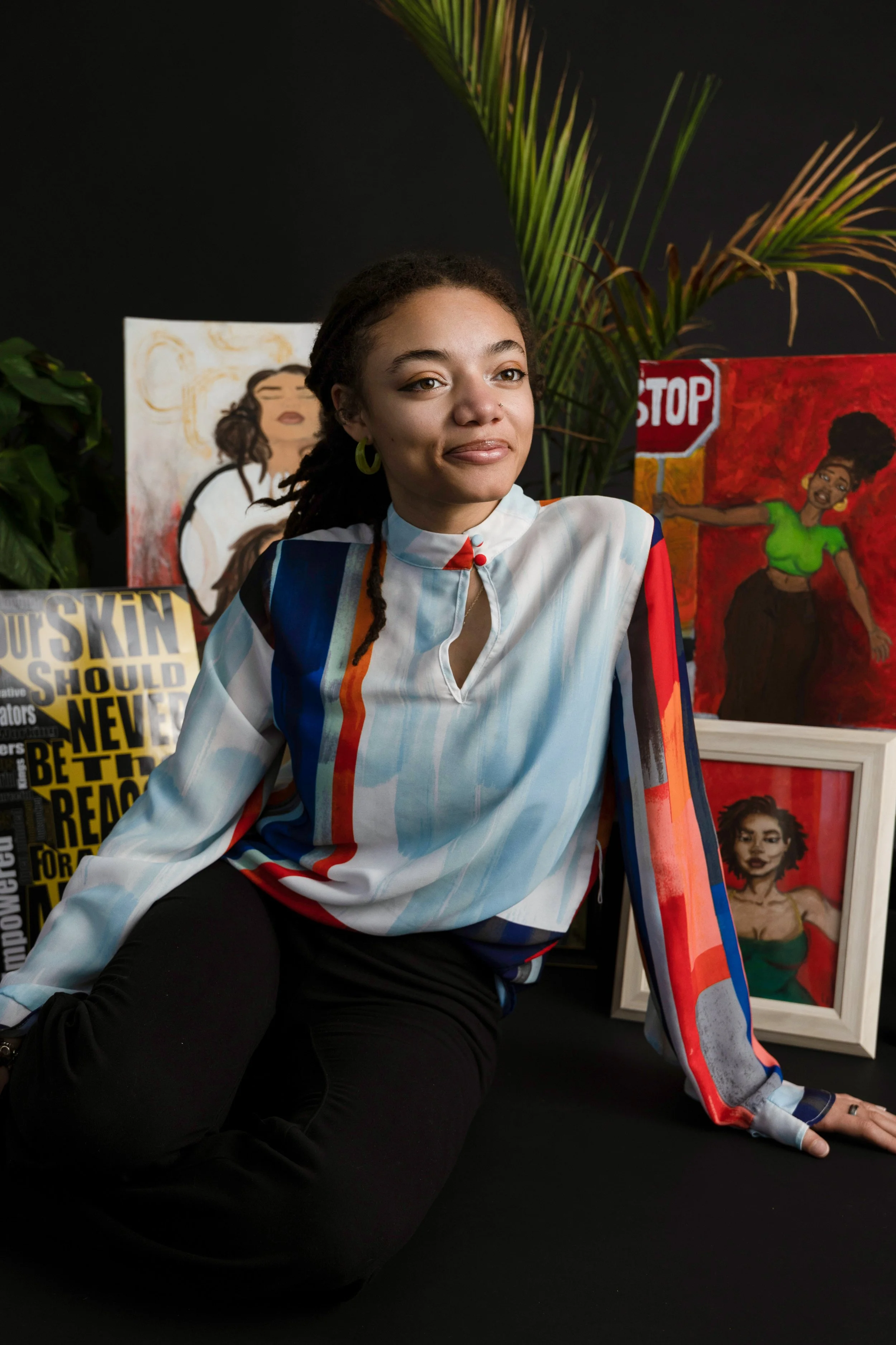 London B (LBL) sitting in front of her paintings, wearing hoop earrings sitting on the floor amongst plants, wearing a multicolored blouse with a high neckline and black pants.