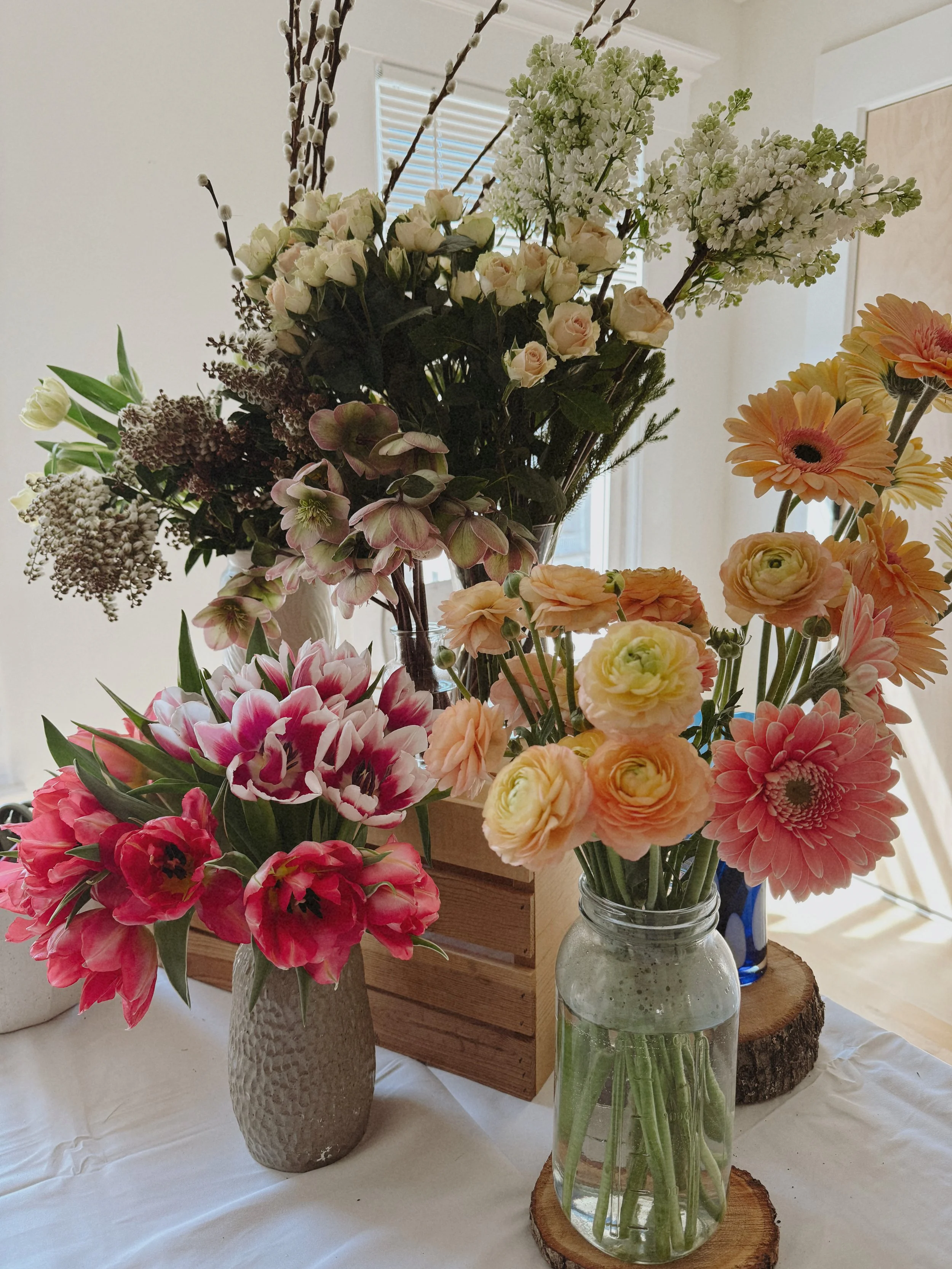 Arrangement of various fresh flowers in vases on a table, with a window in the background.