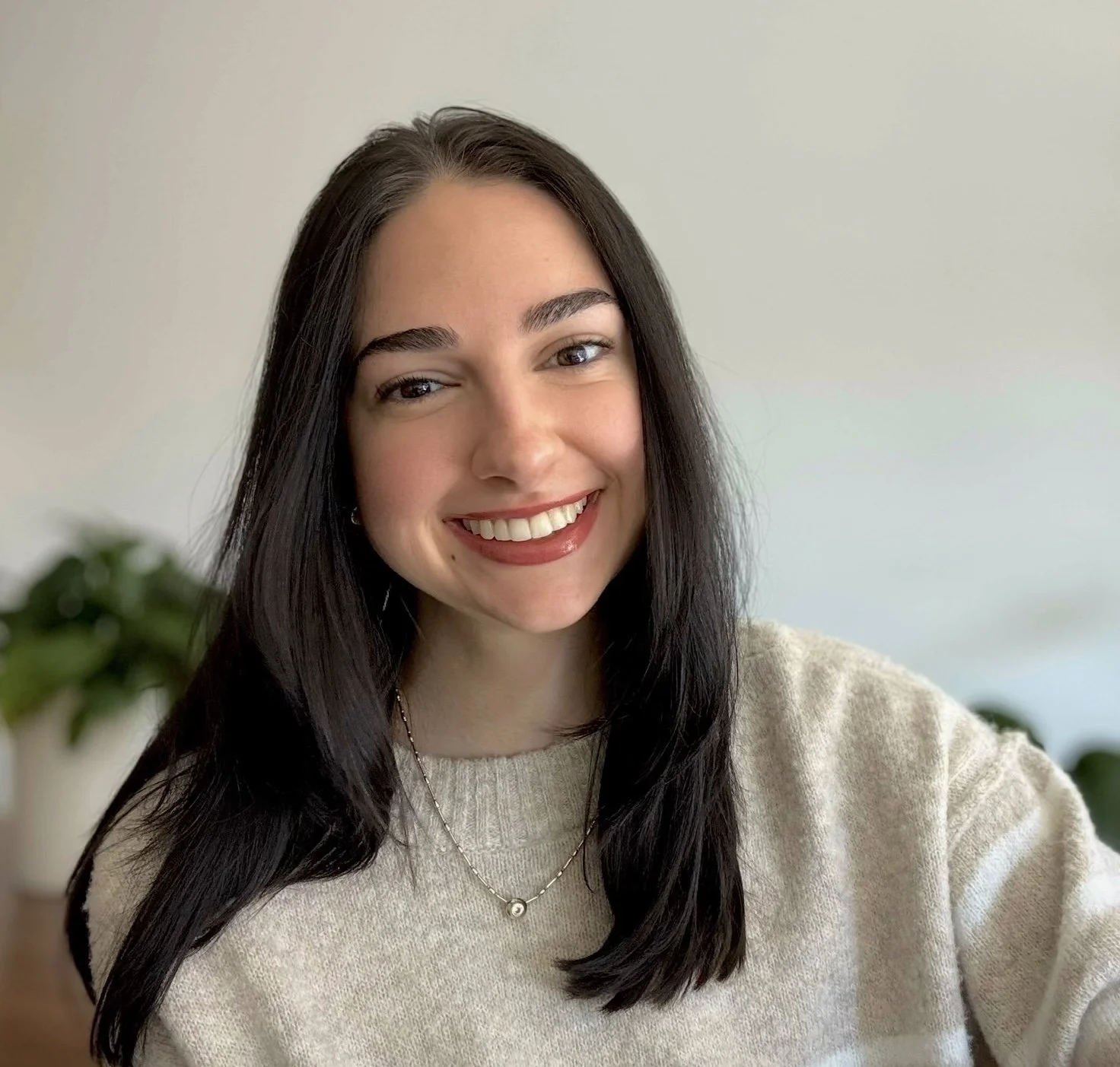 A young woman with long dark hair smiling in a beige sweater, with plants in the background.
