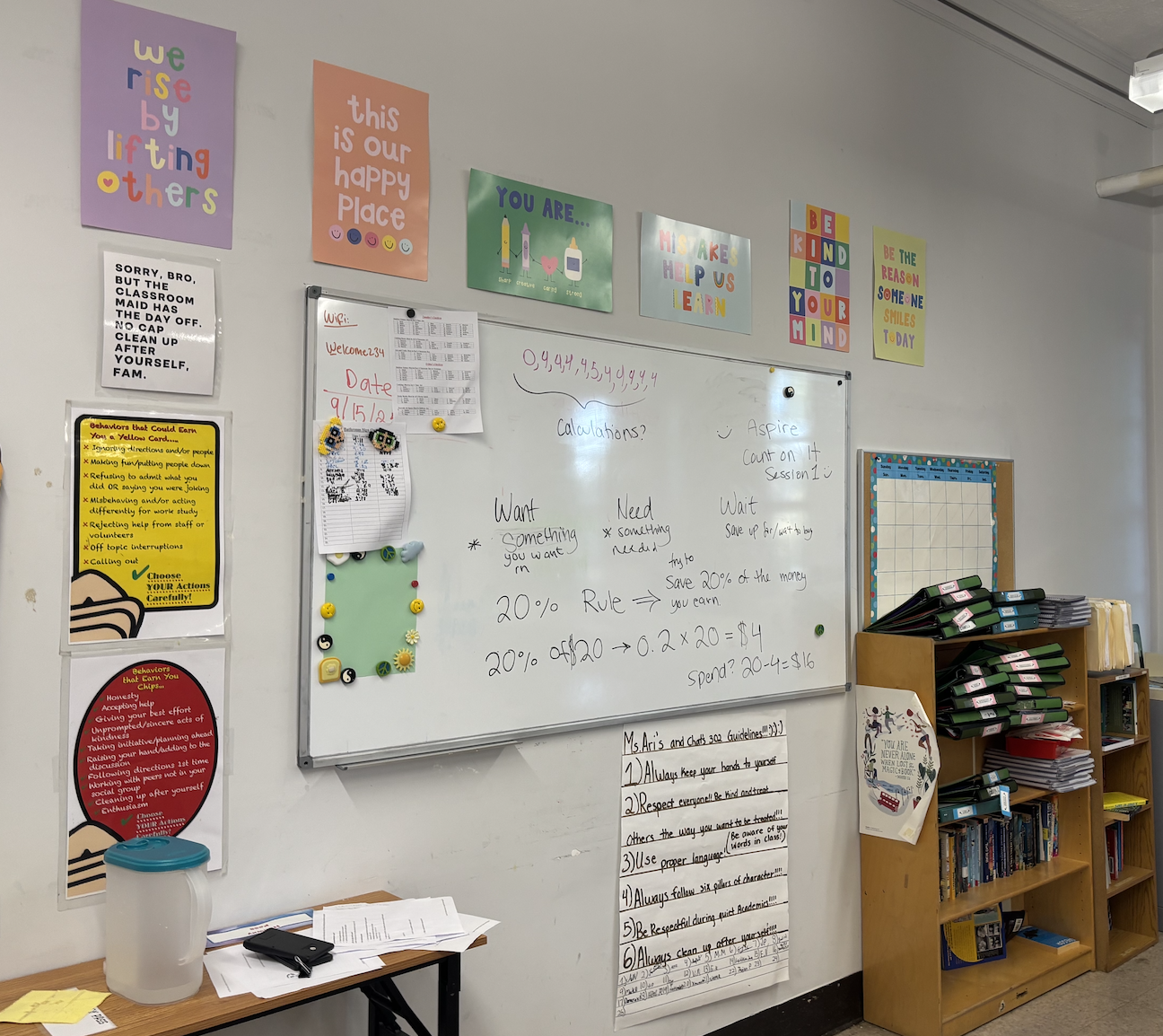 Classroom wall with colorful motivational posters, a whiteboard with handwritten notes about calculations, and a wooden bookshelf filled with folders and books.