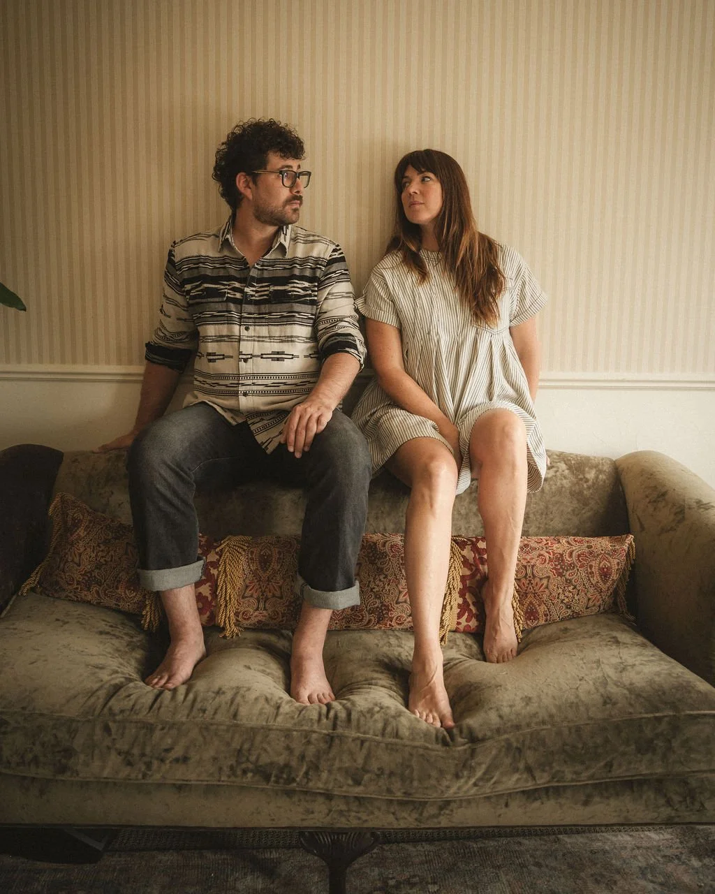 A man and woman sitting on a vintage sofa against a striped wallpaper wall, looking at each other.