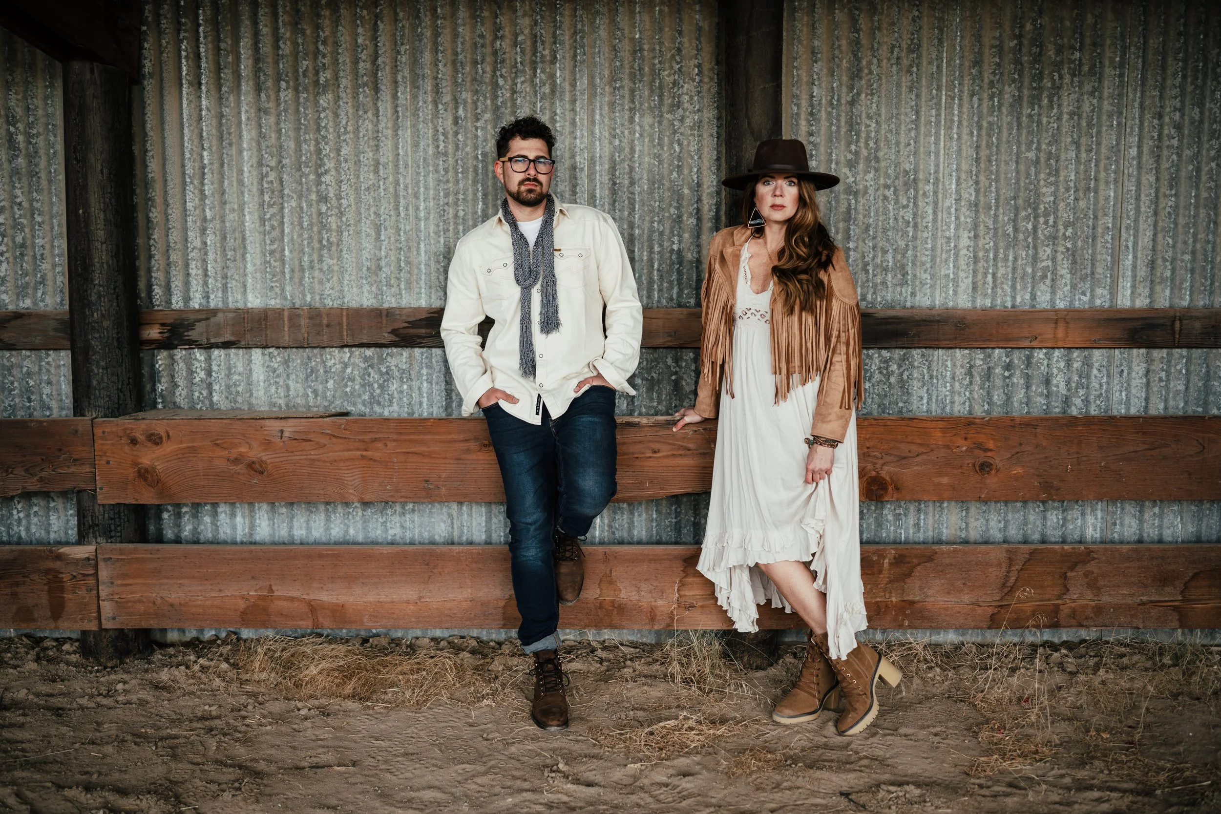 A man and woman standing in front of a wooden and corrugated metal wall. The man is wearing a white shirt, jeans, and boots, with glasses and a scarf. The woman is wearing a long white dress, a fringed jacket, a wide-brimmed hat, and boots, with long