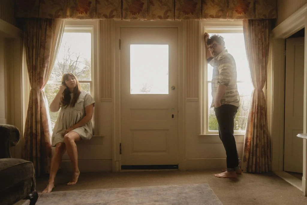 Two women in a vintage-style room with floral curtains, one sitting on a window sill and the other standing near a window, both looking pensive.