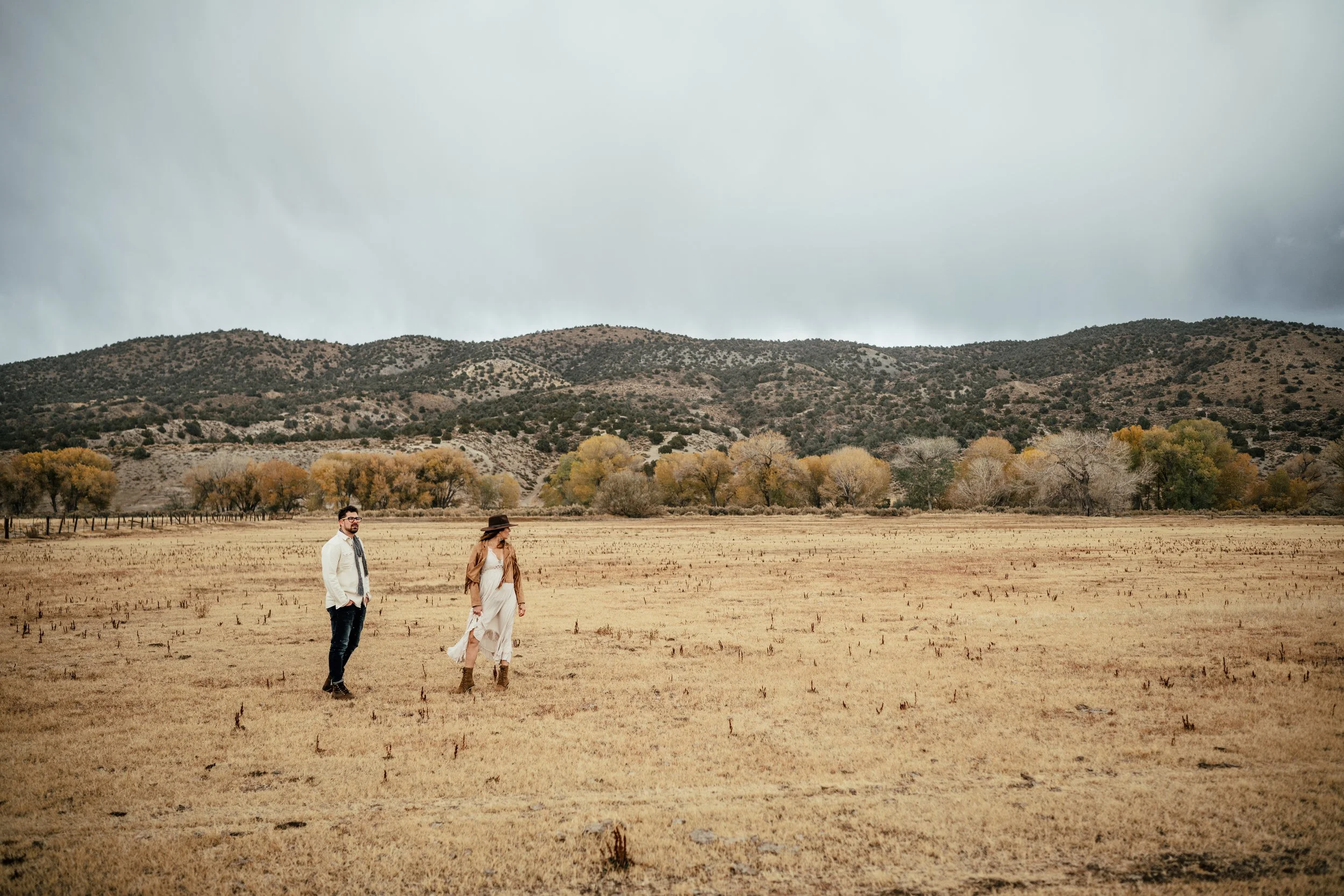 A man and woman standing in a dry field with mountains and trees in the background under cloudy sky.