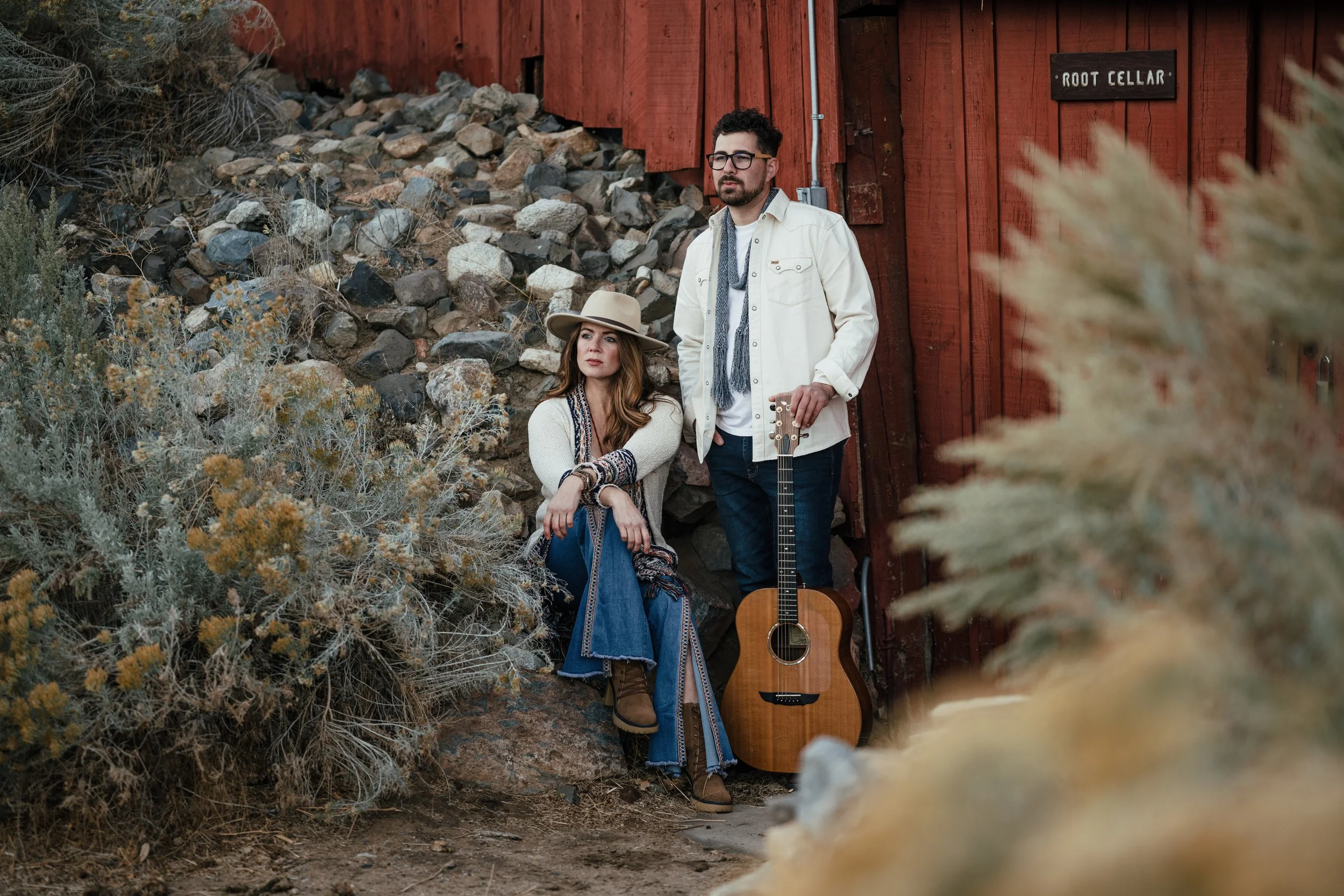 A man and woman outdoors near a red wooden building, surrounded by bushes and rocks. The woman is sitting on a rock, wearing a hat and casual clothing, while the man is standing holding a guitar, dressed in a white jacket and glasses.