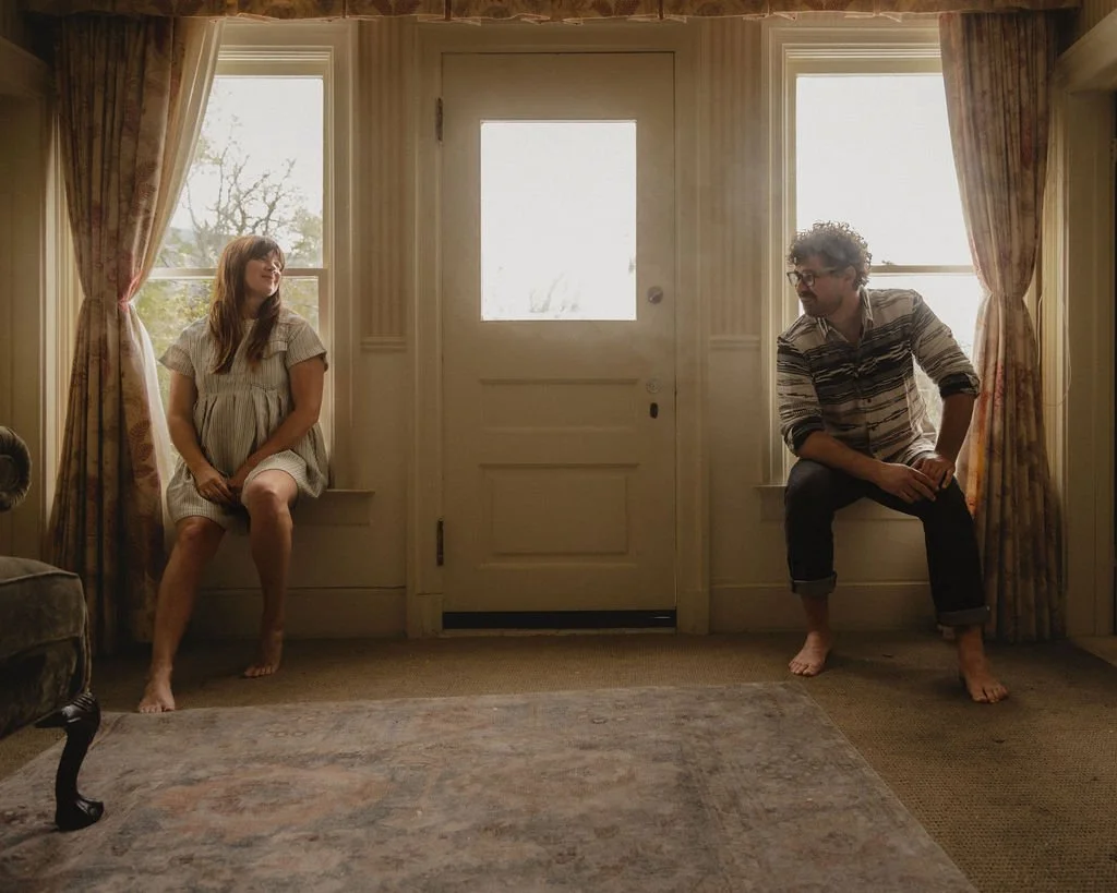 A young woman and a young man sitting on window seats on either side of a closed door in a living room, with sunlight coming through the windows.