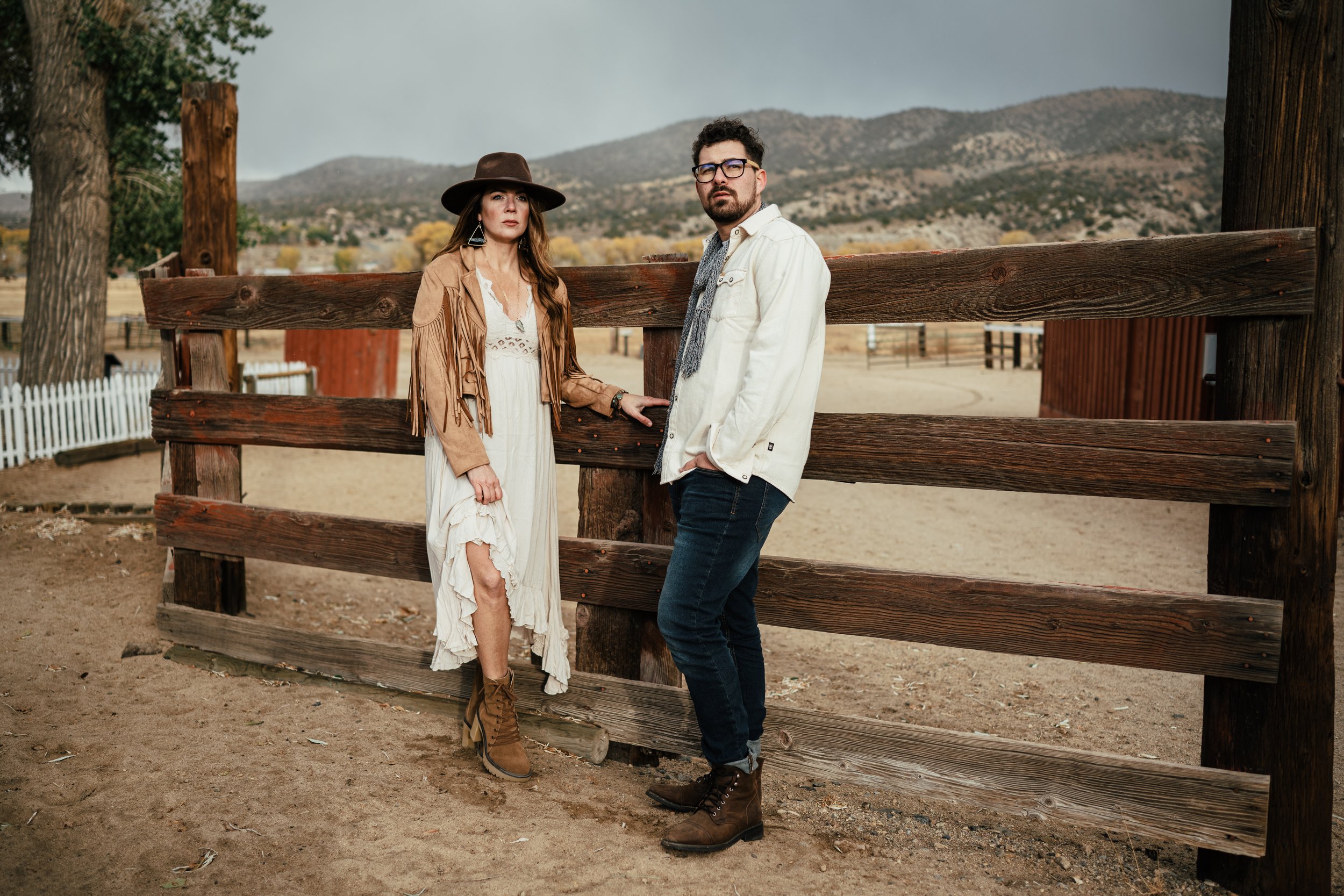 A woman and a man standing on a dirt ground in front of a wooden fence in a rural area with mountains in the background. The woman is wearing a wide-brimmed hat, a fringe jacket, and a white dress. The man is wearing glasses, a white jacket, dark jea