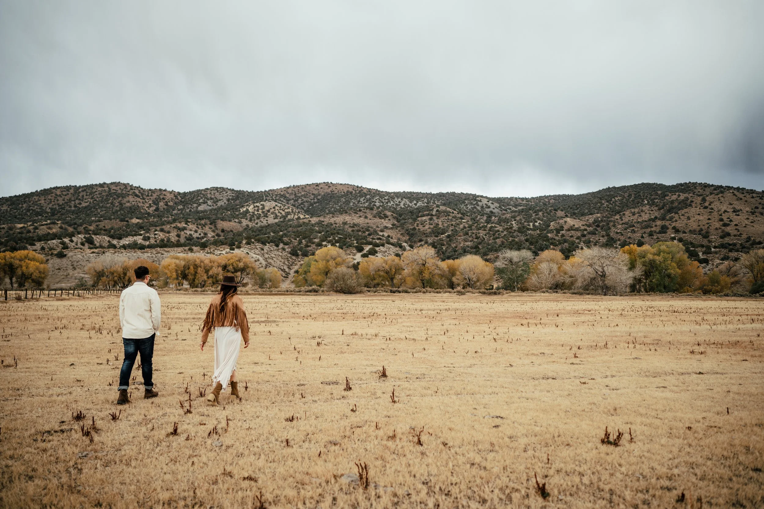 Two people walking across a dry field with mountains in the background and overcast skies.