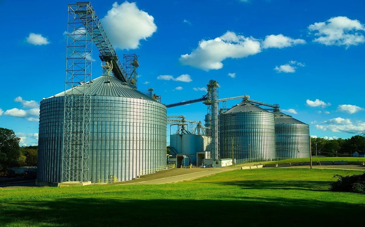 A farm with large metal grain silos under a bright blue sky with white clouds.