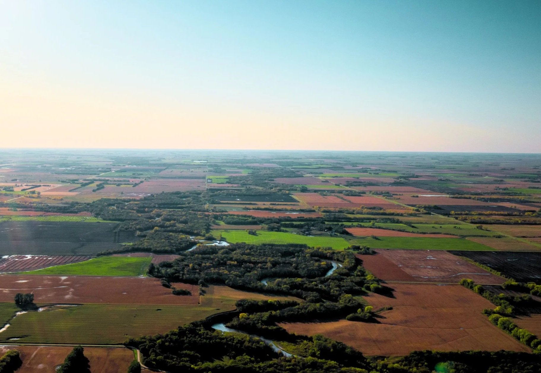 Aerial view of farmland with fields, patches of green and brown, trees, a winding river, and a clear sky