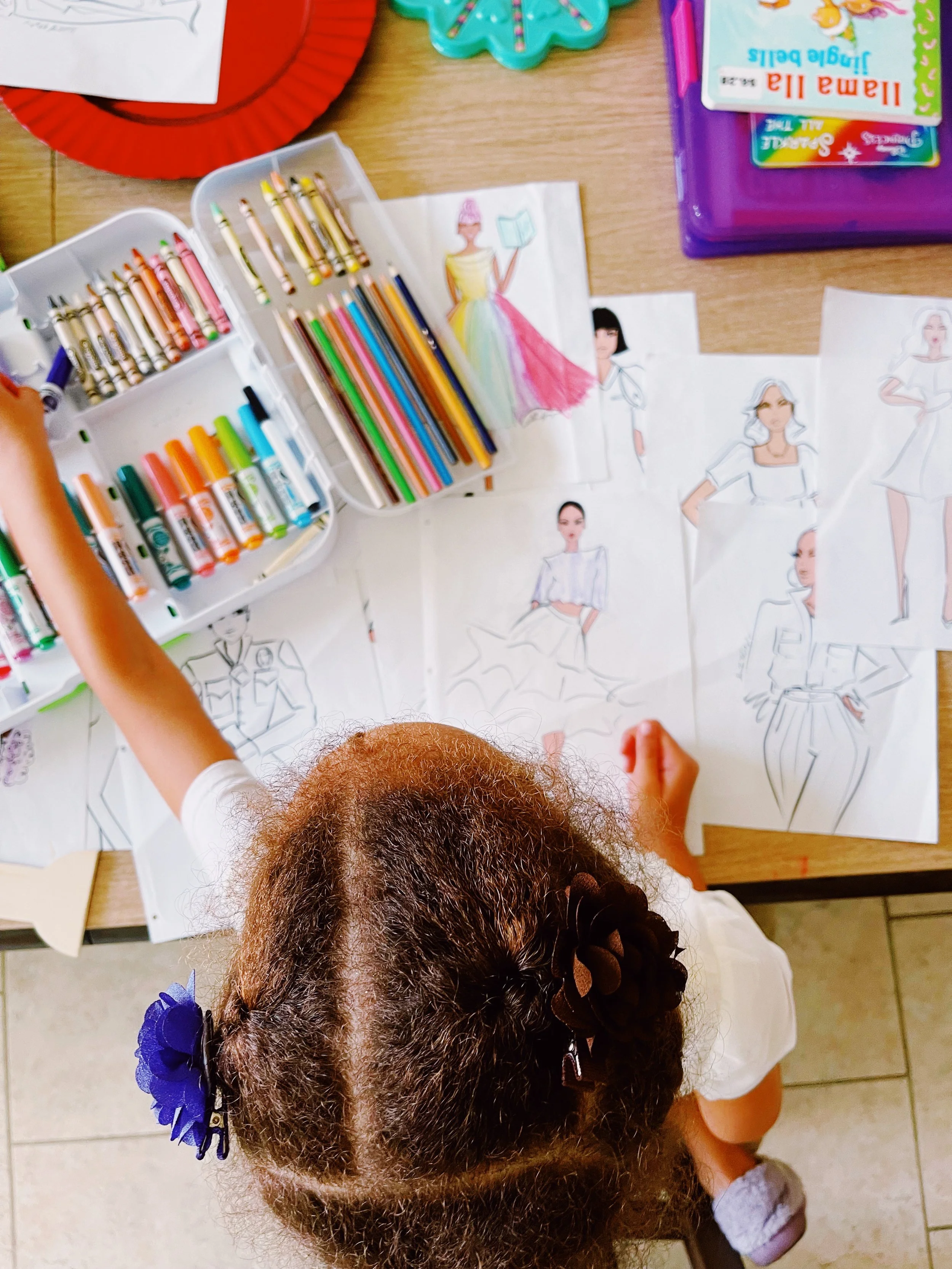 A top-down view of a young girl drawing and coloring fashion sketches at a table. The table is scattered with various colored pencils, crayons, and fashion illustration drawings.