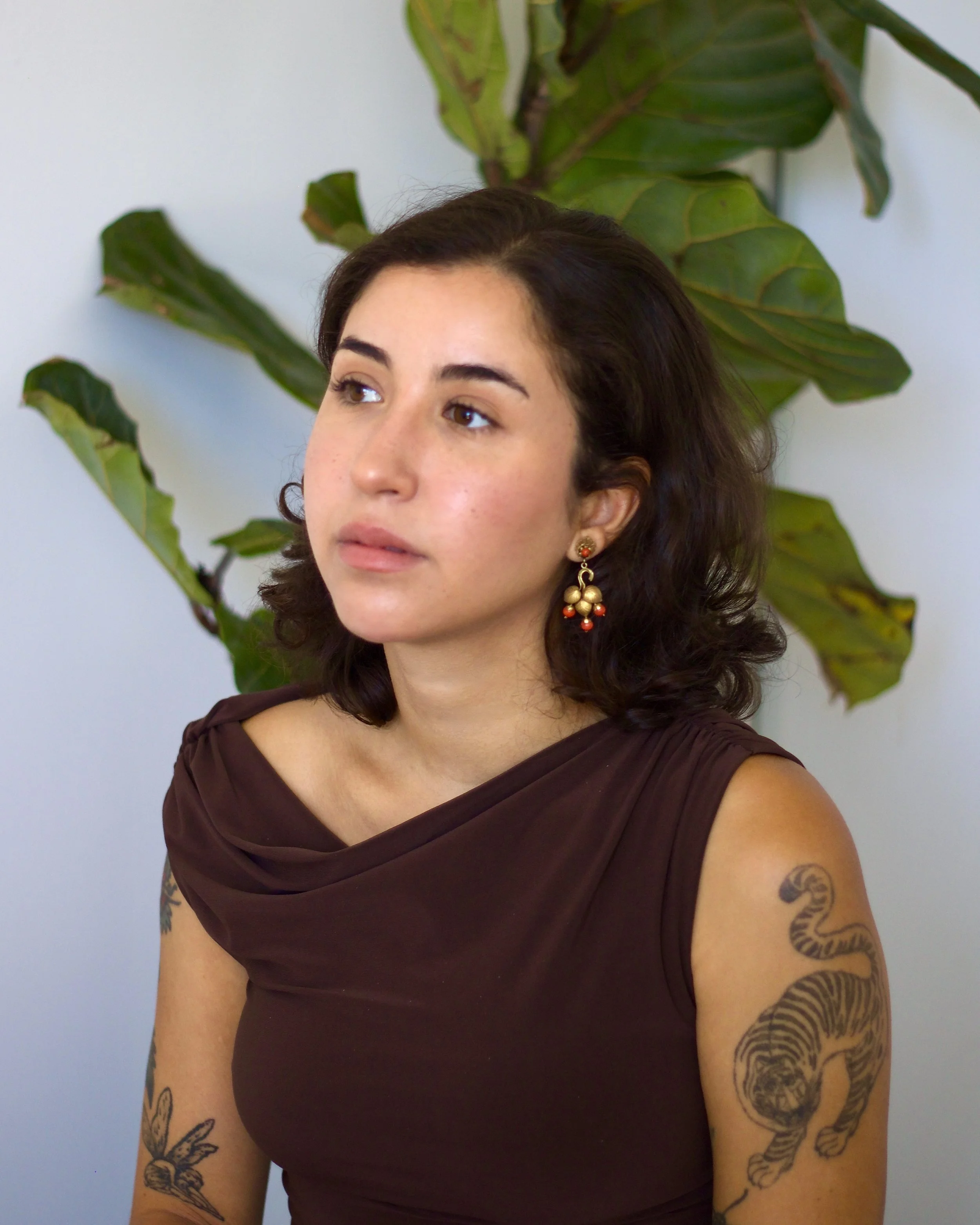 A woman with dark hair and earrings, wearing a brown top, sitting in front of a large green plant with broad leaves.