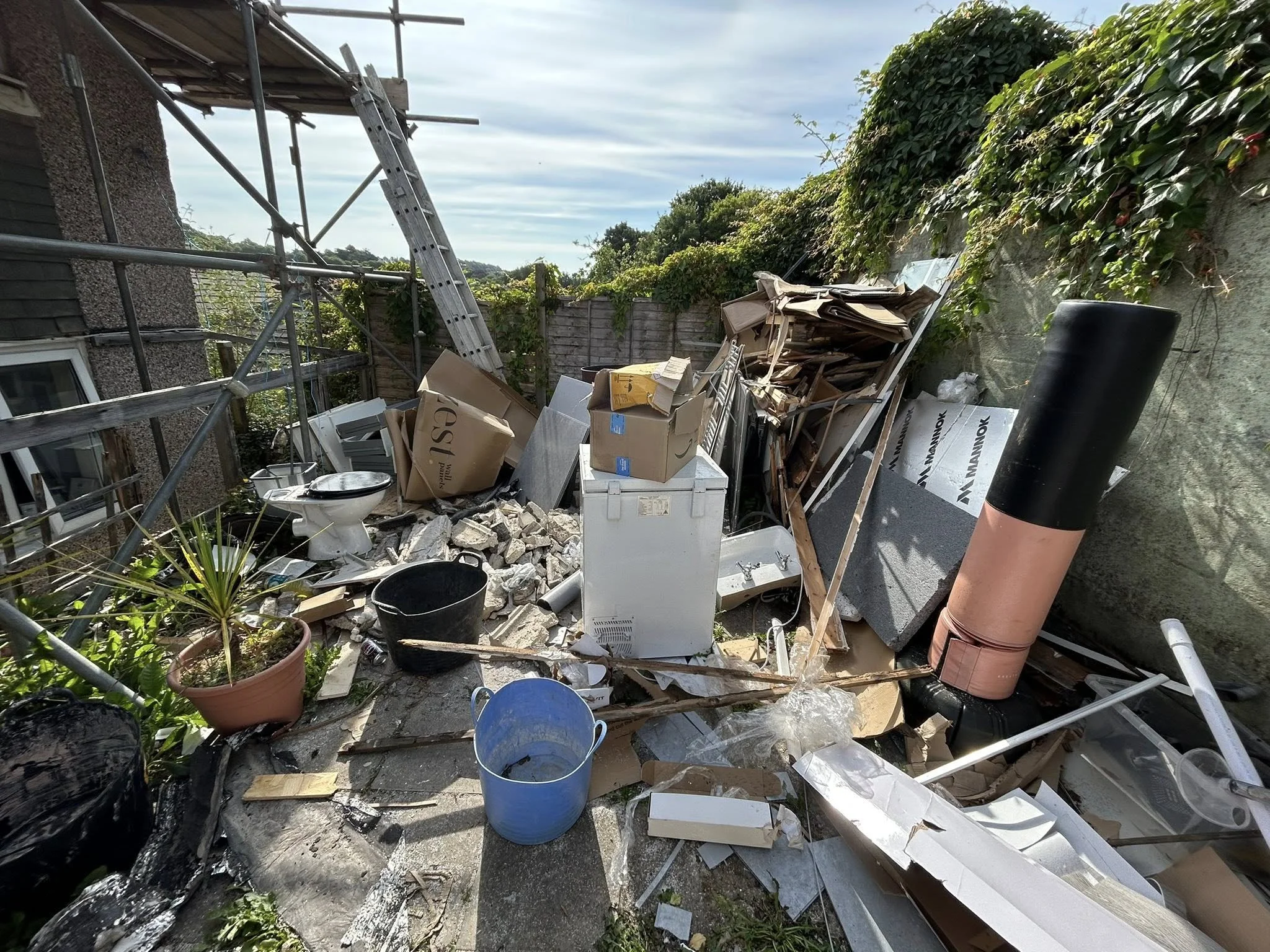 Messy backyard debris including cardboard boxes, construction materials, a potted plant, black plastic bucket, washing machine, and a chimney pipe, with a wooden fence and greenery in the background.
