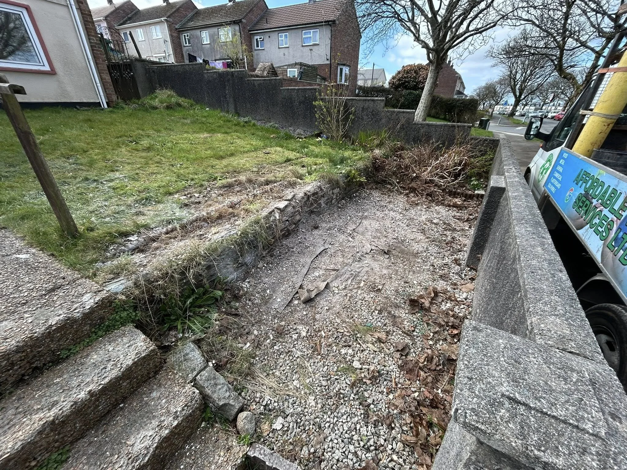 A small area of a sidewalk and stairs with damaged ground, some grass, bushes, trees, and a truck parked nearby.
