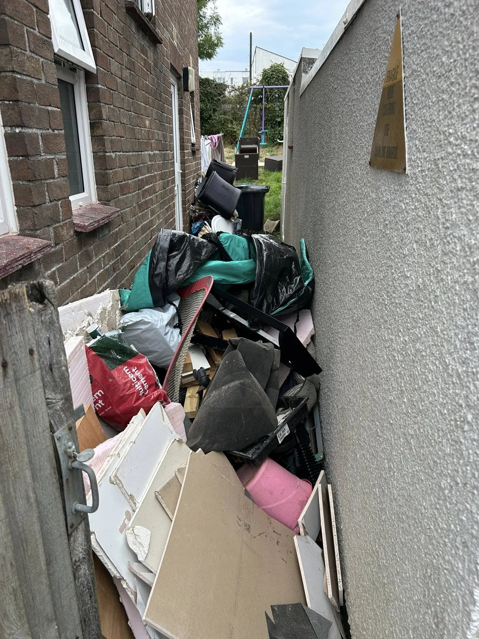 Cluttered backyard alley with trash bags, discarded furniture, and miscellaneous debris.