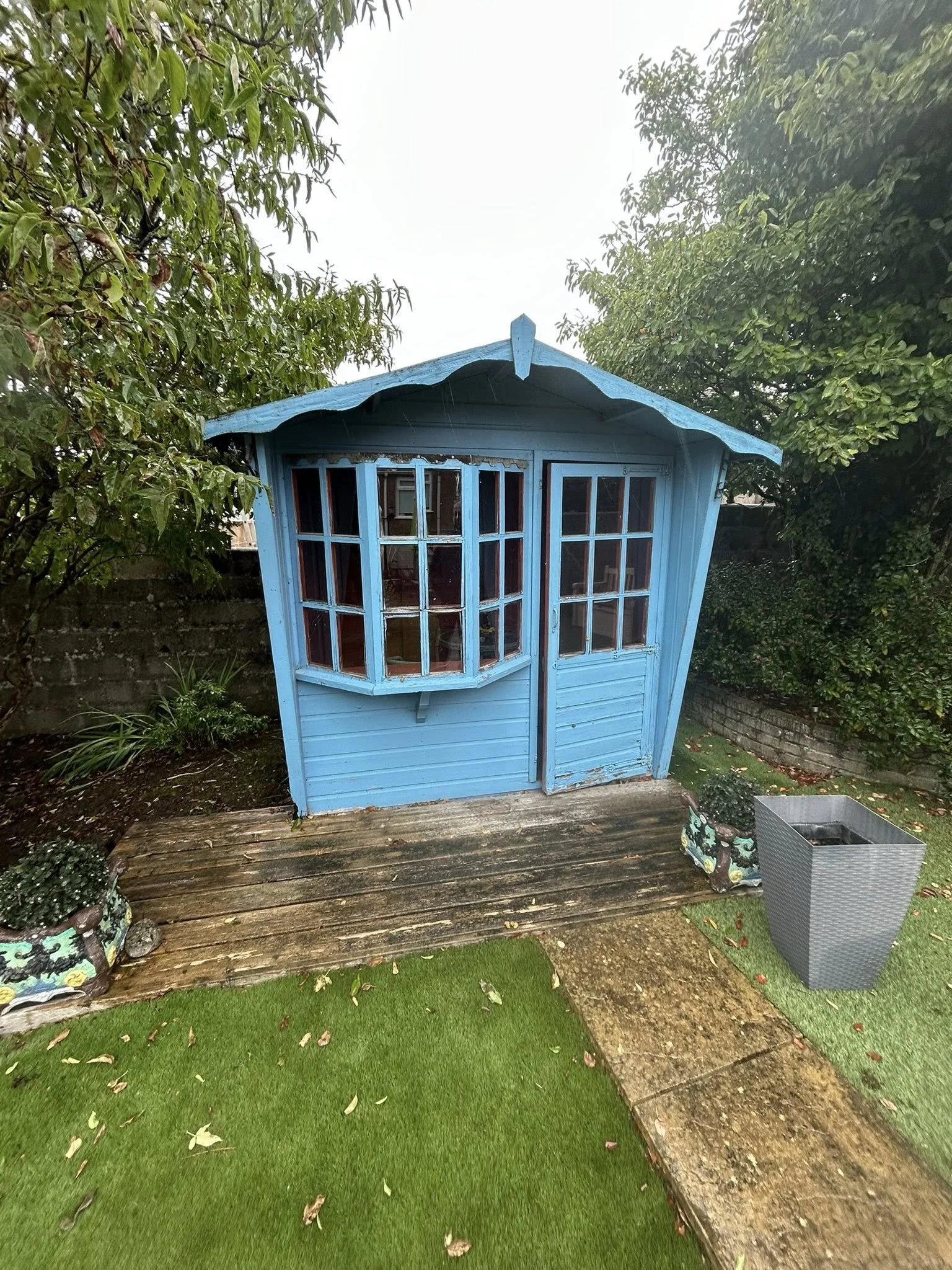 Small blue wooden shed with large windows, a door, and a sloped roof, situated on a wooden deck in a backyard with green grass, trees, and plants.