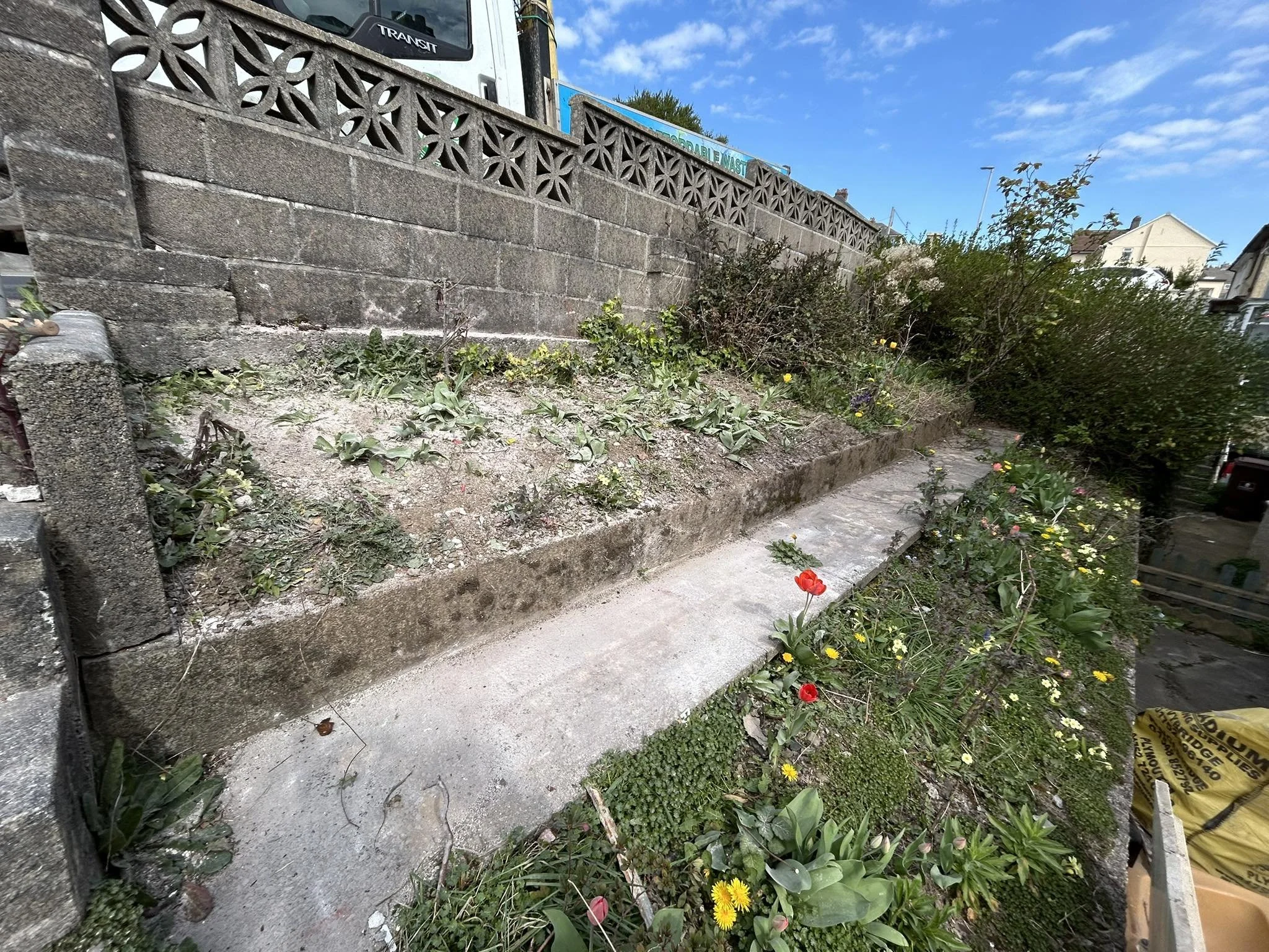 A sidewalk with a garden bed on a hillside, bordered by a brick wall and trucks in the background, under a blue sky with scattered clouds.
