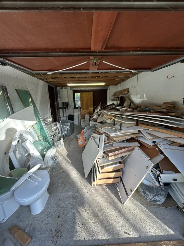 Garage with construction materials, cabinets, and bathroom fixtures including a toilet, stacked on the floor and shelves.