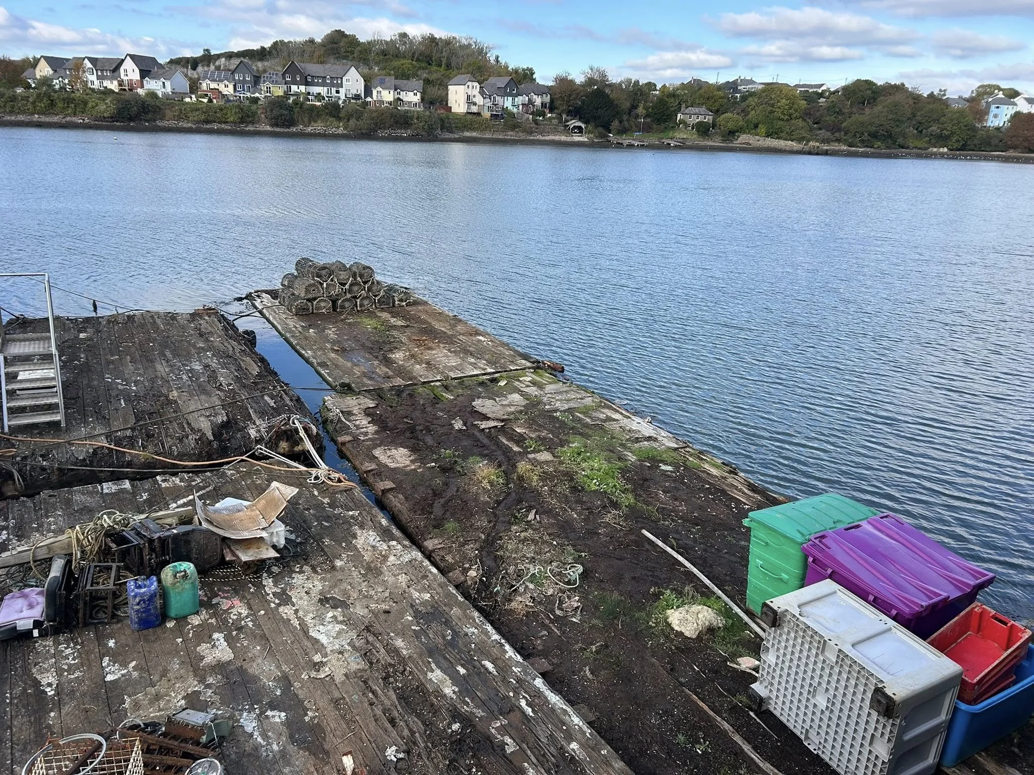 A weathered wooden dock by a body of water with colorful plastic storage bins and miscellaneous objects on it, and a row of houses across the water.
