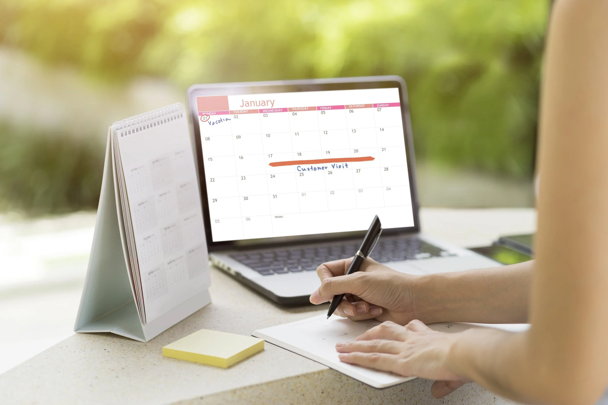 A person writing in a planner at a desk with a laptop and calendar, with a blurred outdoor background.