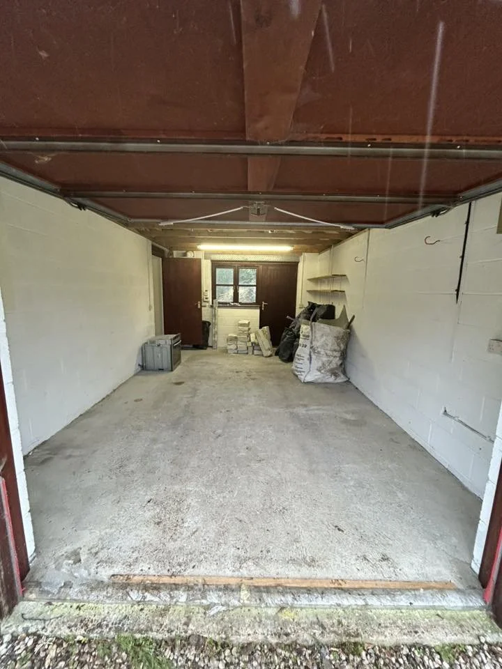Empty garage with concrete floor, white walls, and a red garage door open to the outside. Items like bags, boxes, and a small cabinet are inside near the back wall.