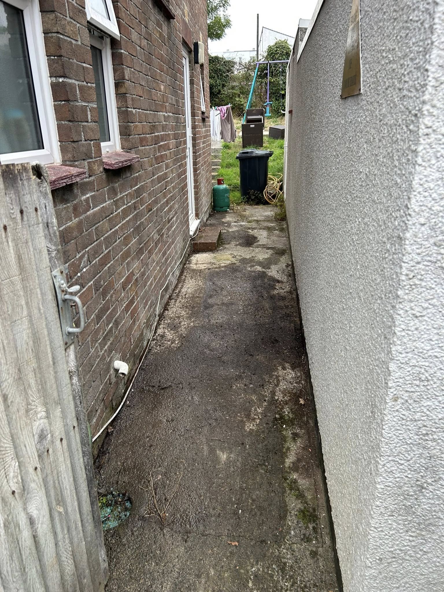A narrow alleyway between two houses, one with brick walls and the other with a white textured wall. The ground is concrete with some dirt patches. At the end of the alley, there is a grassy yard with a children's swing set, a trash can, a propane ta