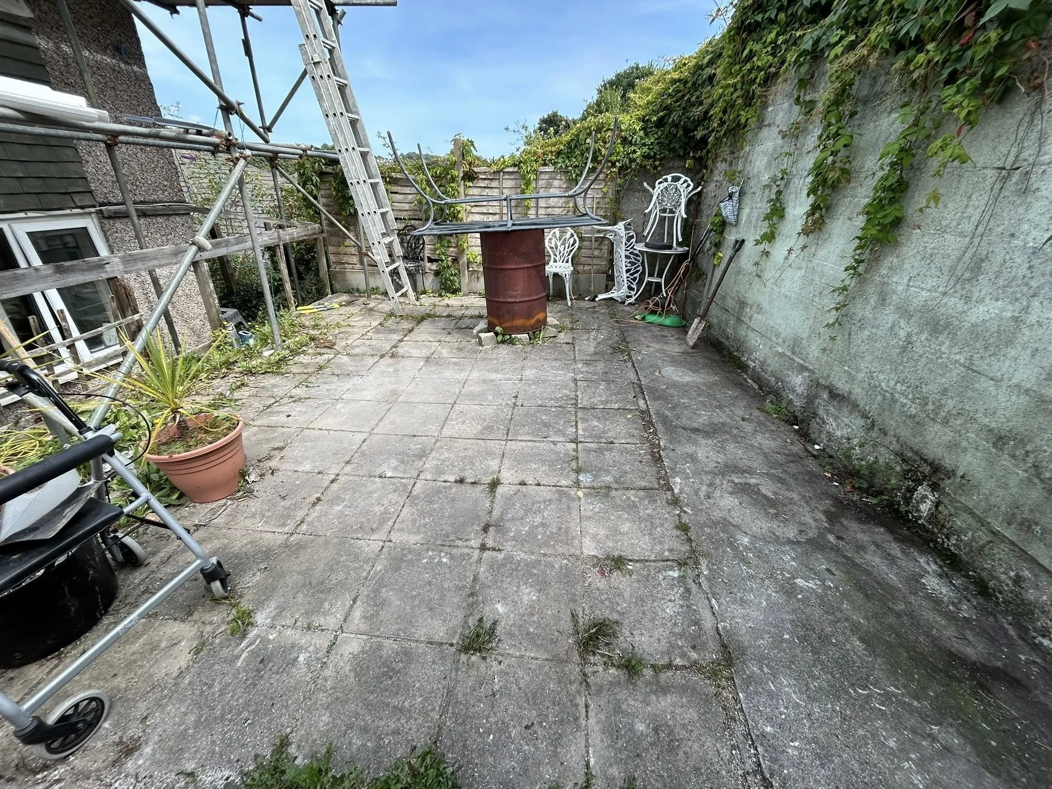 A small outdoor patio with aged concrete pavers, surrounded by a weathered wall and wooden fence. There is a rusty barrel used as a table, white metal chairs, a potted plant on a small table, and gardening tools leaning against the wall. Scaffolding 