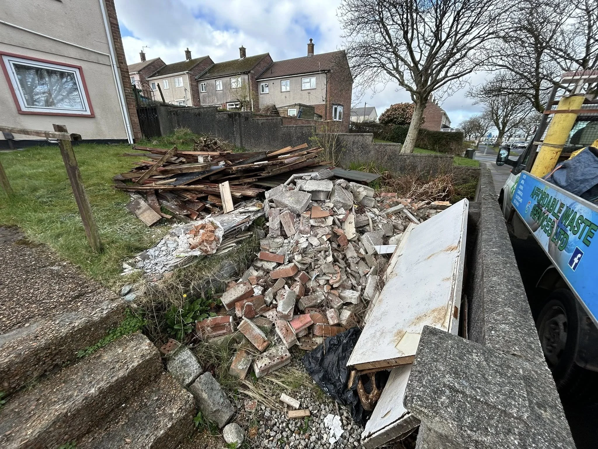 Pile of construction debris, including wooden planks, bricks, and a broken white door, next to a sidewalk and a parked waste collection truck.