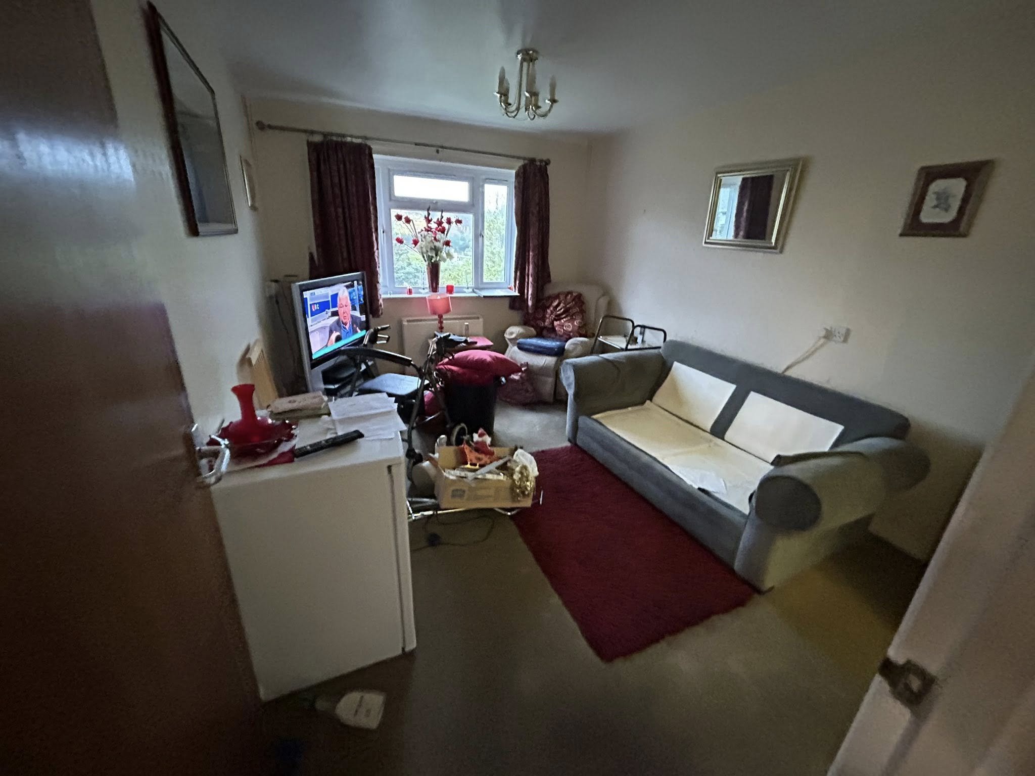 A cluttered living room with a TV on a stand, a gray sofa, a floral armchair, a small red rug, and a window with red curtains. Various items are spread across the room including papers, a red vase, a wheelchair, and a lamp with a red shade.
