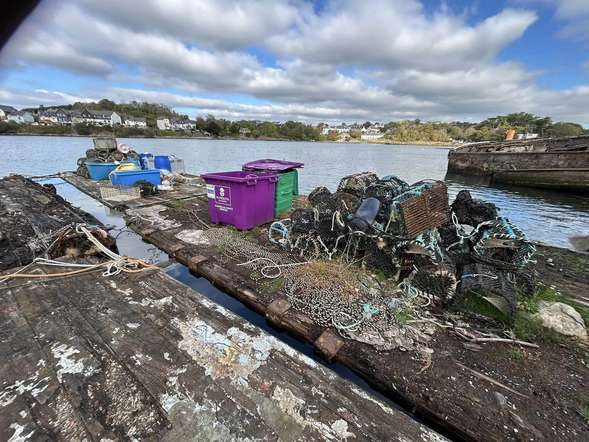 Fishing nets, plastic containers, and trash on a wooden dock by a body of water with houses and trees in the background.