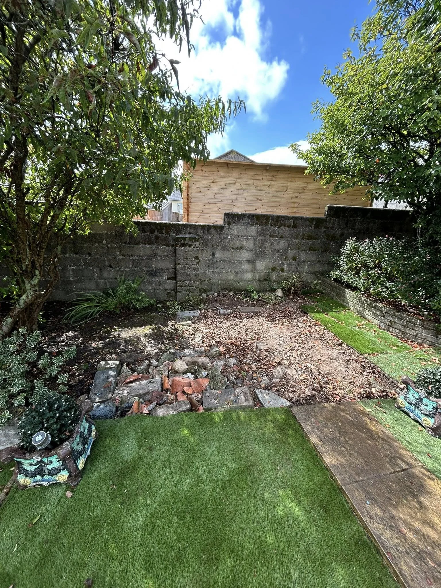 A backyard with artificial grass, a small stone pathway, and a dirt area with rocks and leaves, enclosed by a concrete wall and surrounded by trees and bushes, under a partly cloudy blue sky.