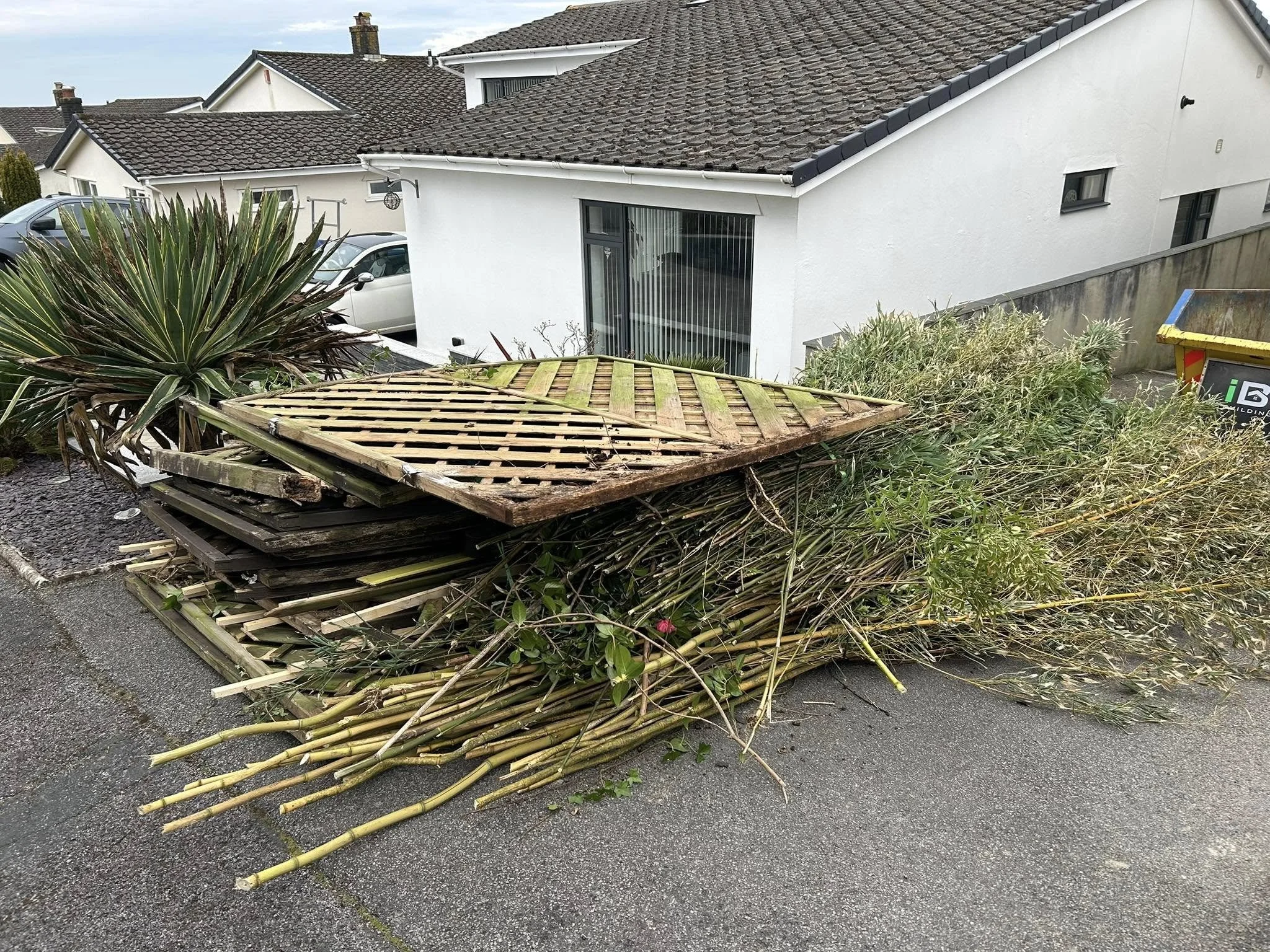 Pile of old wooden pallets and cut bamboo sticks on a driveway in front of a white house.
